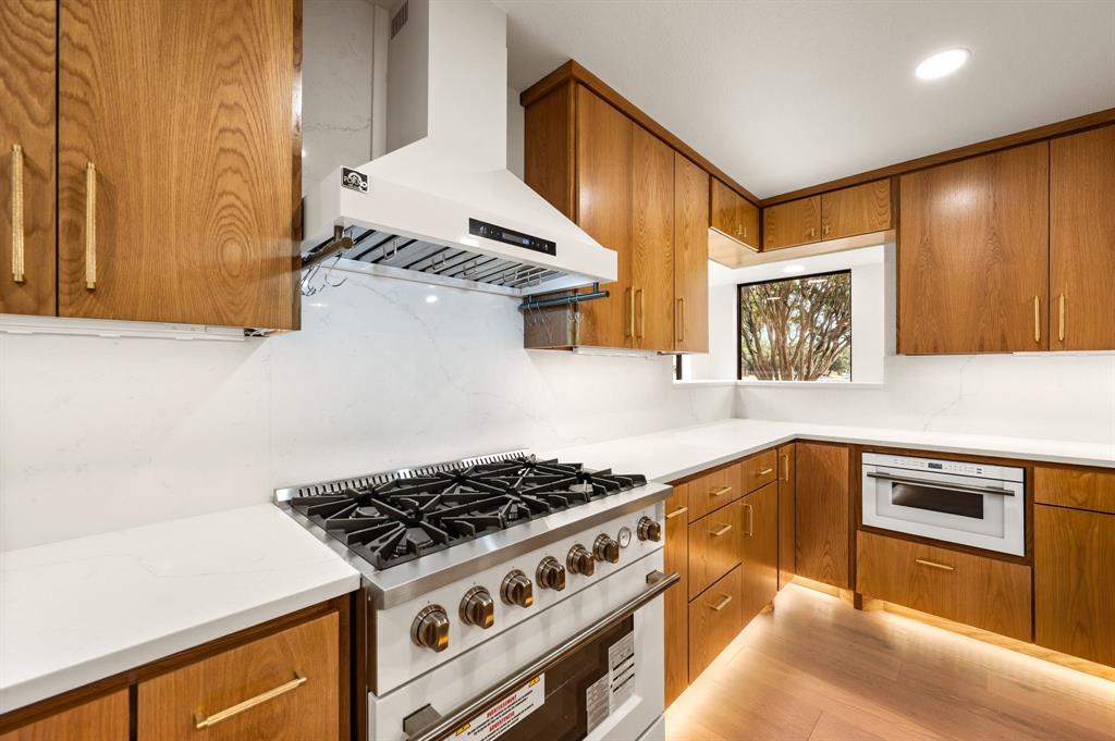6403 Genstar Lane Dallas, TX 75252 - Photo 10 of 34 Gourmet kitchen featuring a stainless steel gas range with a white vent hood, wood-finish cabinetry with brass hardware, solid surface countertops, a built-in microwave, and wood-finish flooring with under-cabinet lighting