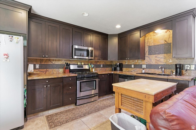 6237 West 4600th Street South Hooper, UT 84315 - Photo 27 of 48 Kitchen featuring stainless steel appliances, dark wood finish cabinets, light stone countertops, light tile patterned floors, and tasteful backsplash