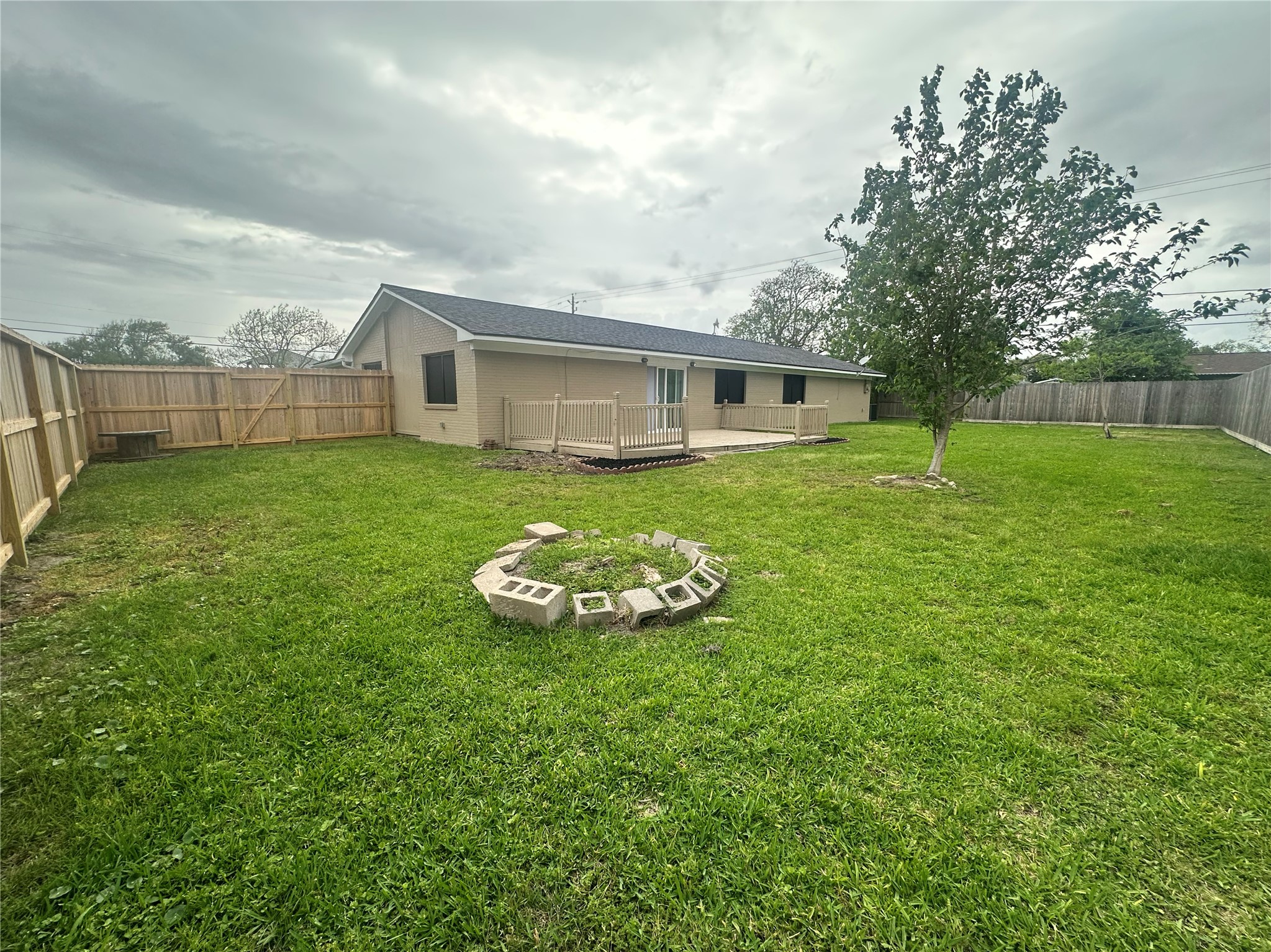 130 Bay Ridge Drive Baytown, TX 77523 - Photo 18 of 20 The photo showcases a spacious backyard with a grassy lawn, a small tree, and a circular arrangement of bricks. The house features a beige exterior with a fenced perimeter for privacy, and a deck area near sliding glass doors. Ideal for outdoor activities and gatherings.
