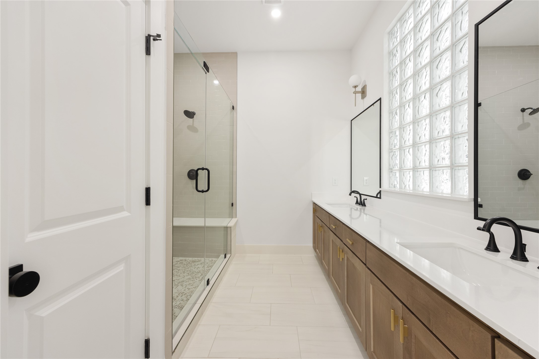 2121 Hoskins Drive, Unit C Houston, TX 77080 - Photo 26 of 34 Spa-inspired primary bath with a long dual-sink vanity, quartz-style countertop, warm cabinetry, black fixtures, brass-toned hardware, glass block window, and a separate glass-enclosed shower. The bright neutral palette, clean lines, and generous counter space create a polished retreat with everyday functionality.