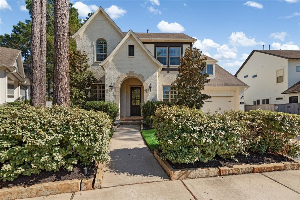 This picturesque two-story brick home features a classic arched entryway, charming dormer windows, and a well-maintained front yard with mature trees and shrubs. The attached garage provides convenient parking.