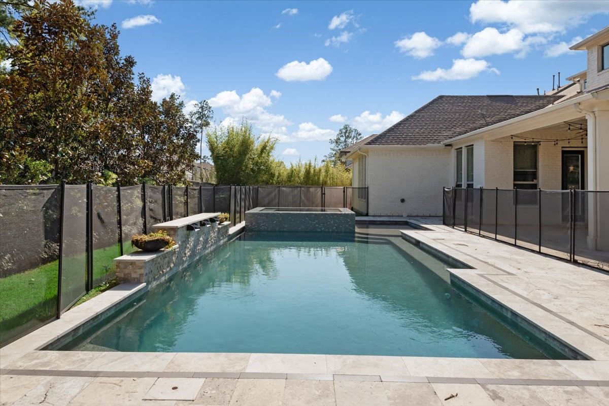 155 Rockwell Park Spring, TX 77389 - Photo 44 of 50 This photo showcases a modern backyard with a spacious rectangular pool, surrounded by a safety fence. The area features a stone deck, a small waterfall, and is bordered by lush greenery, creating a private and serene outdoor retreat.