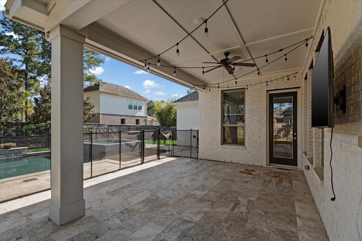 155 Rockwell Park Spring, TX 77389 - Photo 45 of 50 This photo showcases a spacious covered patio with string lights and a ceiling fan, overlooking a pool secured by a safety fence. The area is paved with stone tiles and features a mounted television, making it ideal for outdoor relaxation and entertainment.