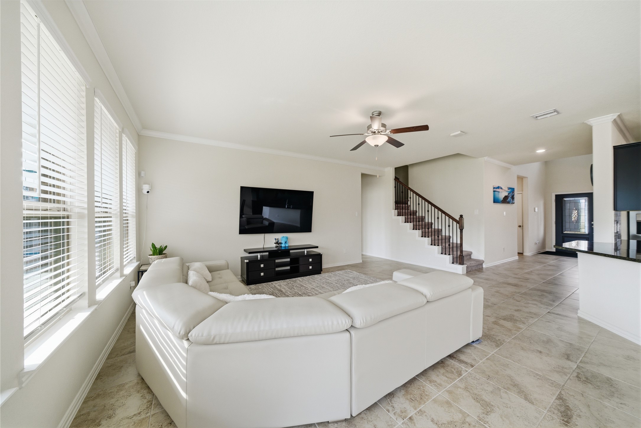 4335 Tawny Timber Drive Spring, TX 77386 - Photo 10 of 42 Bright and spacious living room with large windows, modern tile flooring, a ceiling fan, and an open staircase. It features a white sectional sofa, wall-mounted TV, and easy flow to the kitchen area.
