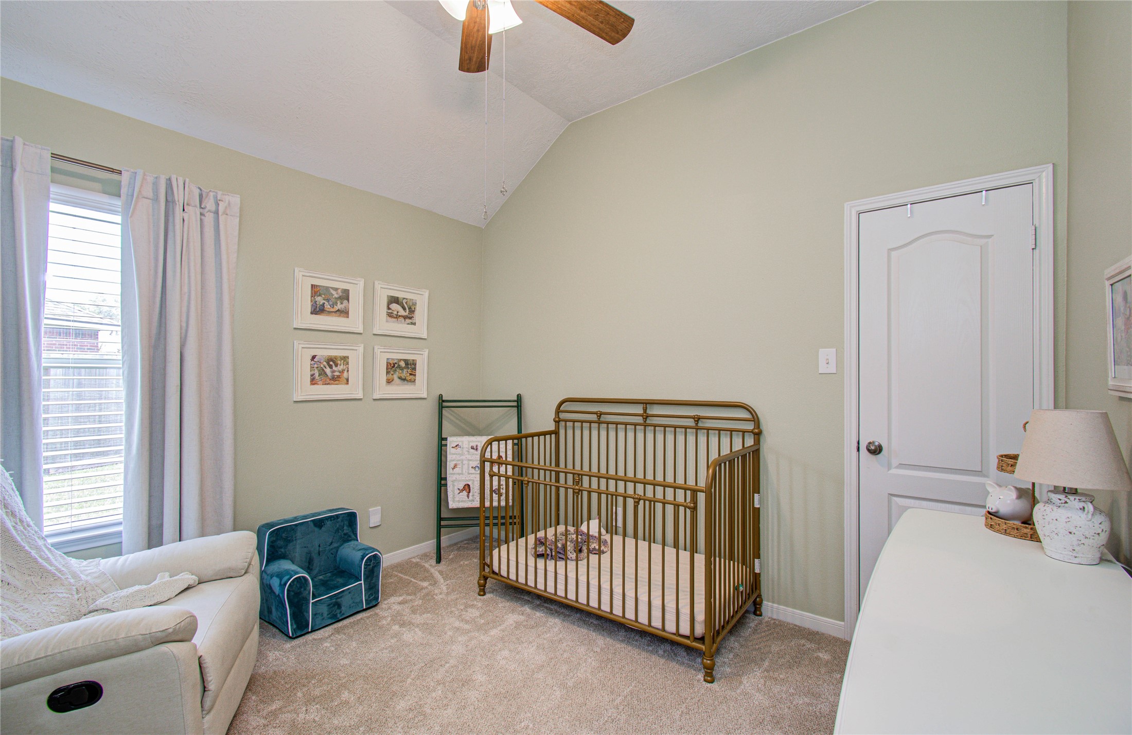 20007 Alcea Court Spring, TX 77379 - Photo 22 of 36 This cozy nursery is adjacent to the additional room/den. It features soft green walls, a vaulted ceiling with a ceiling fan, and a large window for natural light. It includes a crib, a comfortable recliner, and a small bookshelf, creating a welcoming space for a baby.