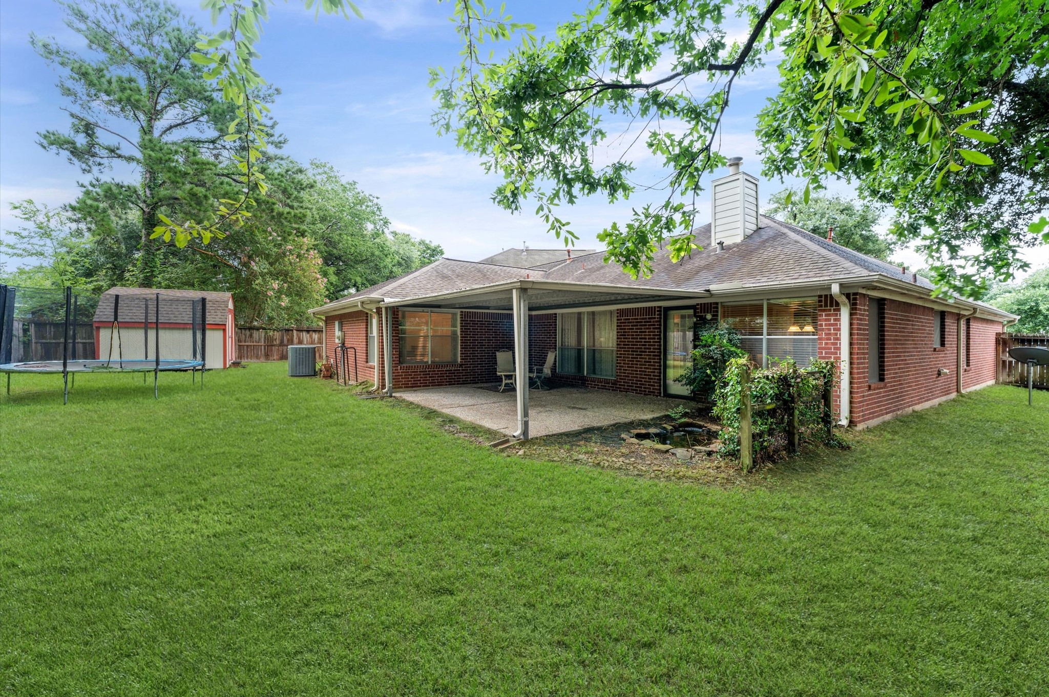 17618 Mill Springs Drive Spring, TX 77379 - Photo 24 of 26 This photo shows a charming one-story brick house with a spacious, well-maintained backyard. The yard features a covered patio, perfect for outdoor relaxation, and a storage shed for extra space. Lush greenery and a trampoline add to the family-friendly appeal.