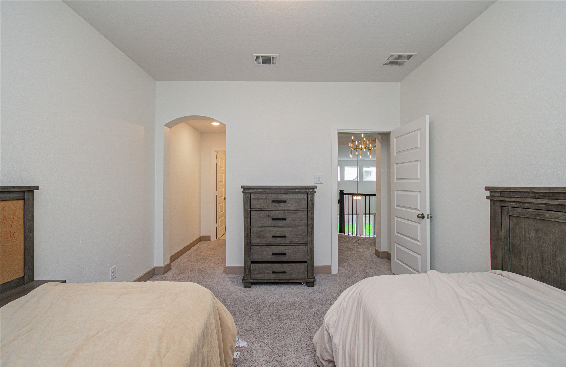 4211 Limestone Terrace Manvel, TX 77578 - Photo 25 of 33 This photo shows a cozy bedroom with neutral walls and carpet, featuring two beds and a dark wood dresser. The room has an arched doorway leading to a hallway and a door opening to a view of a staircase with a chandelier.
