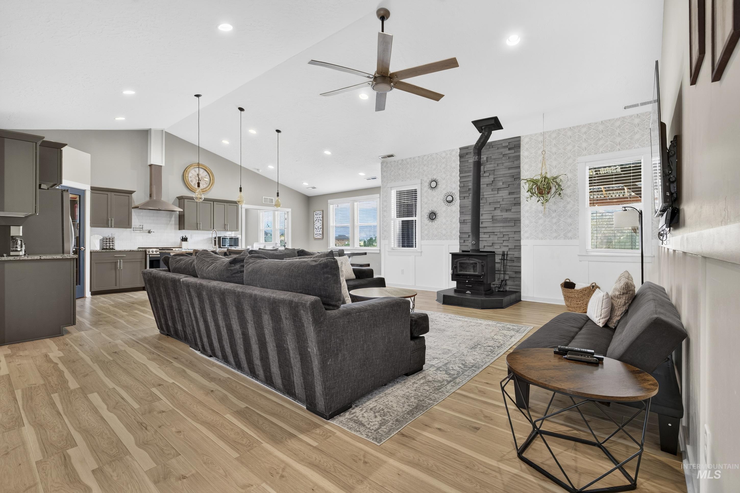 132 Mores Creek Rim Road Boise, ID 83716 - Photo 7 of 44 Open-concept living space featuring vaulted ceilings, wood-finish flooring, a wood-burning stove with a stone surround, and an integrated kitchen with gray cabinetry and pendant lighting