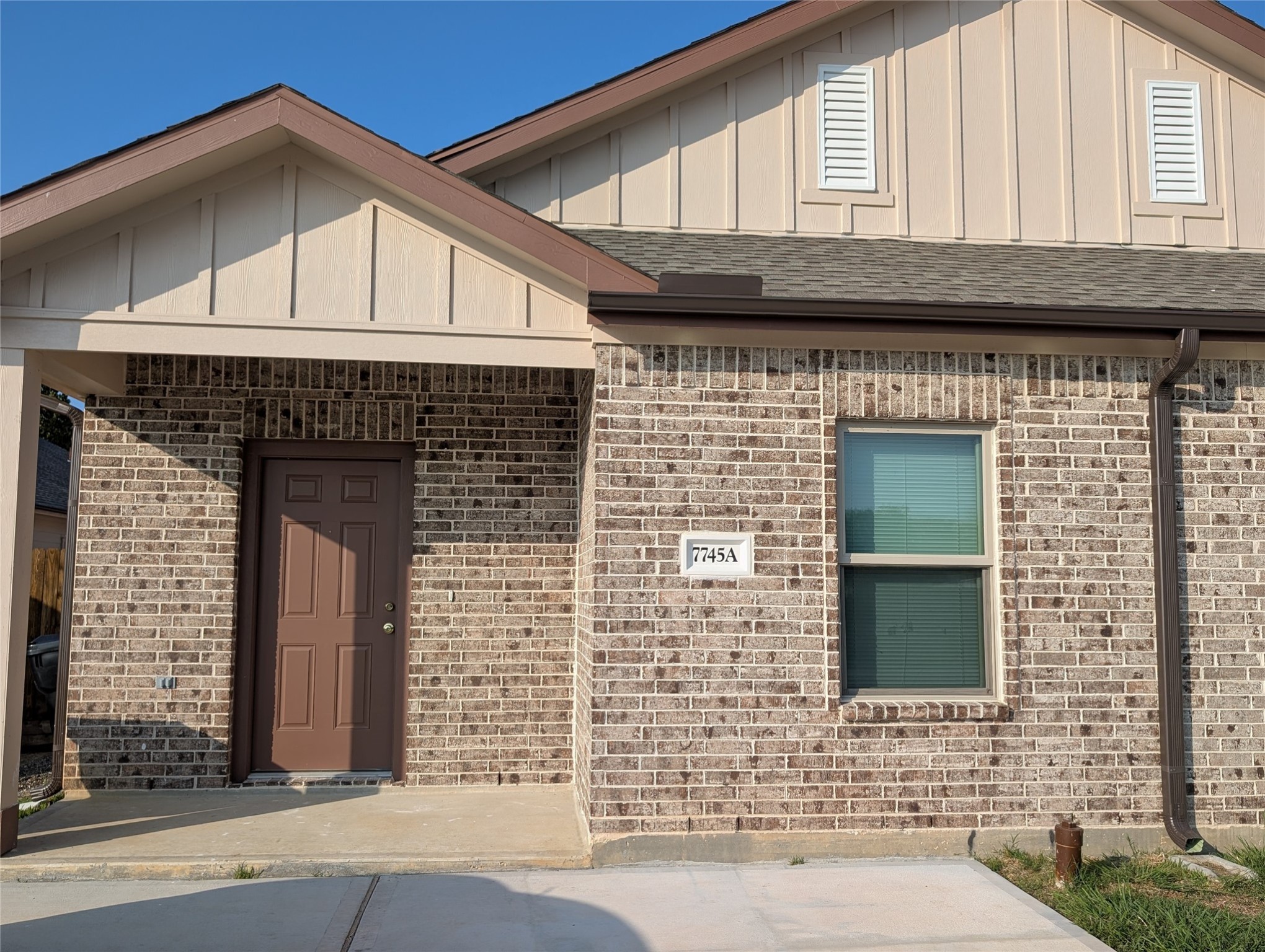 7745 Swiftwater Lane, Unit A Houston, TX 77075 - Photo 2 of 33 This is a close-up shot of the front entry of a new construction home (unit 7745A), showcasing multi-toned brown brick and light tan vertical siding. The entrance is sheltered by a small covered porch with a brown door, alongside a single window.
