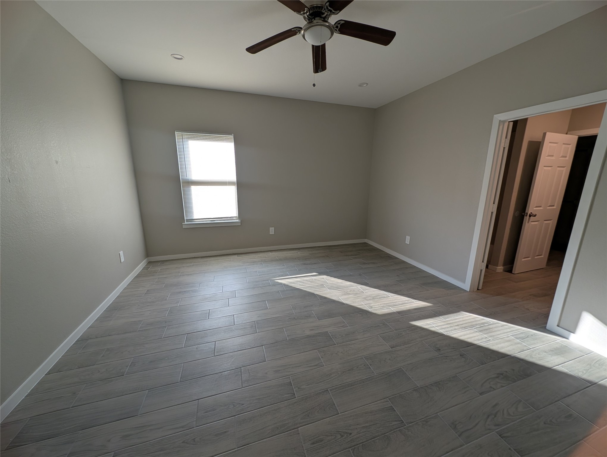 7745 Swiftwater Lane, Unit A Houston, TX 77075 - Photo 26 of 33 This bright and spacious primary bedroom showcases high-quality grey wood-look tile flooring and neutral walls, offering a clean canvas. It receives plenty of natural light from the window and includes a dark wooden ceiling fan with a central light fixture.