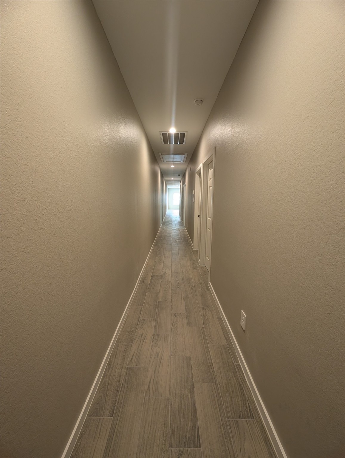 7745 Swiftwater Lane, Unit A Houston, TX 77075 - Photo 29 of 33 This exceptionally long, straight hallway extends from the back to the front of the home, featuring consistent grey wood-look tile flooring and neutral tan walls with bright white trim. The passage is well-lit by recessed ceiling lights and includes a visible air vent.