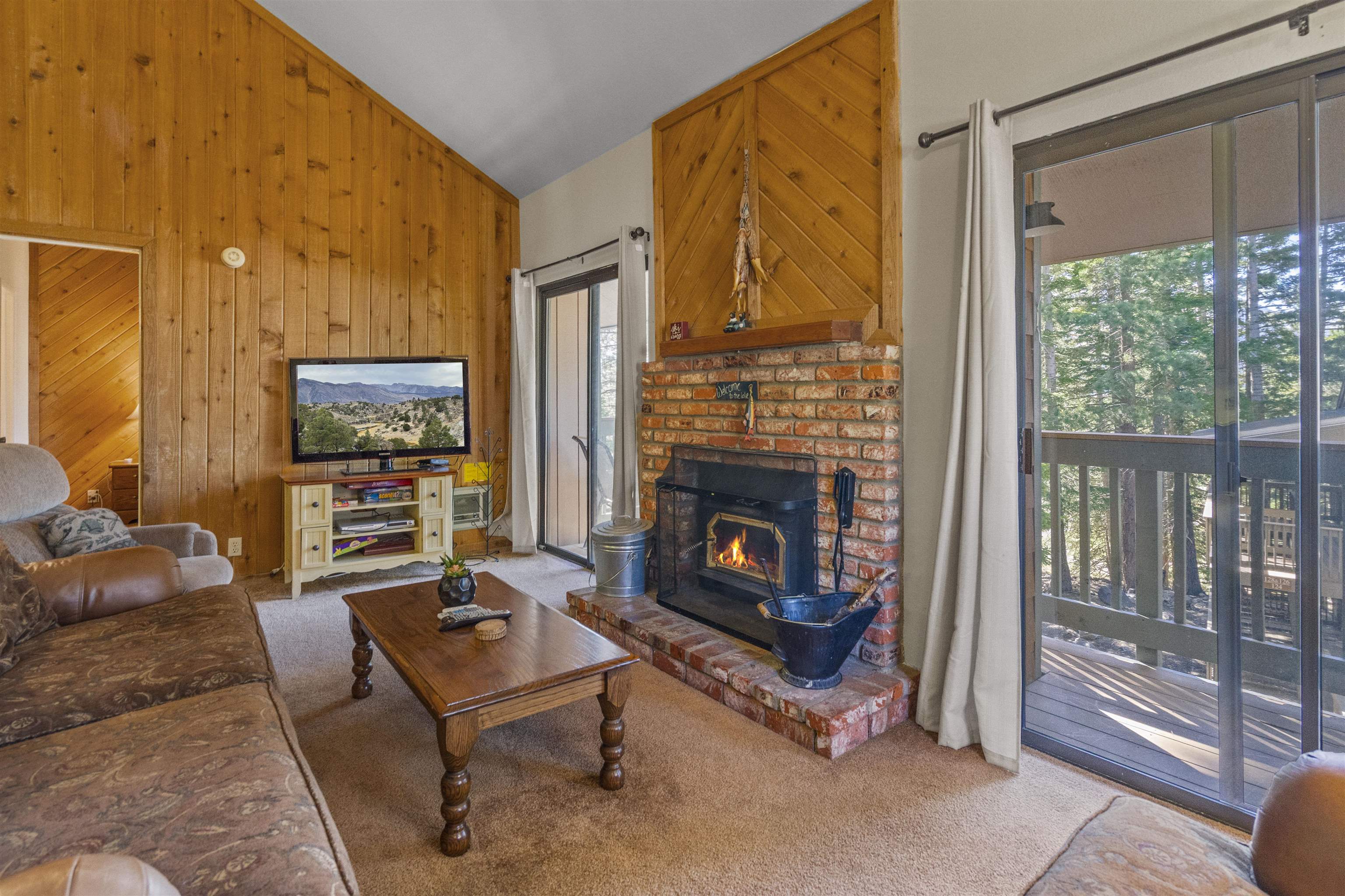 153 Lake Mary Road, Unit 114 Mammoth Lakes, CA 93546 - Photo 2 of 22 Living area with vaulted ceilings featuring wood paneling, a brick fireplace with a wood-burning insert, and sliding glass doors opening to an exterior deck