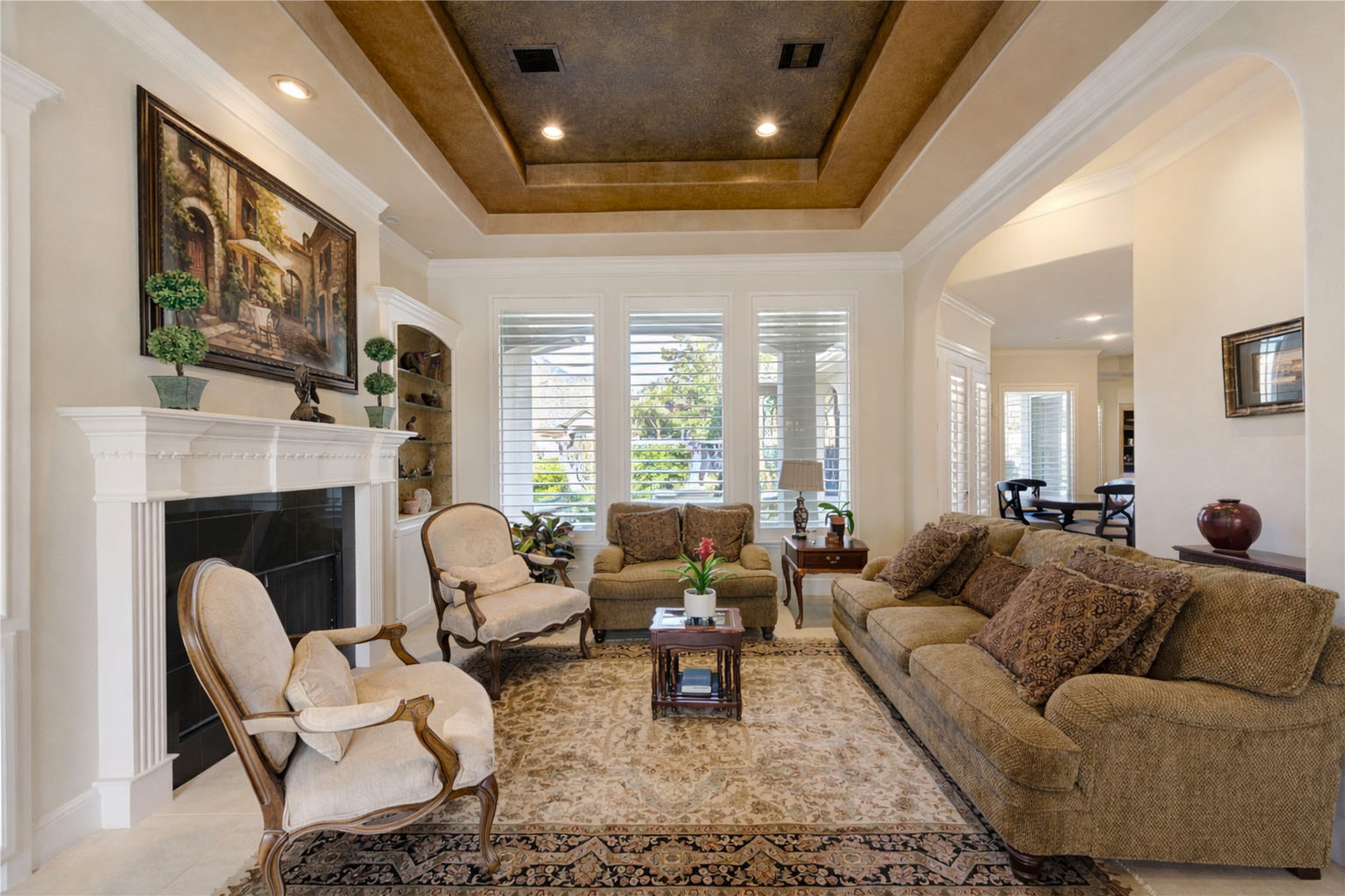 16618 Victoria Falls Lane Spring, TX 77379 - Photo 6 of 49 This living room features a warm, inviting ambiance with a coffered ceiling and recessed lighting. A classic fireplace is accented by tasteful décor, while large windows with plantation shutters allow plenty of natural light. The room is furnished with plush seating and a patterned area rug, creating a cozy yet sophisticated atmosphere.