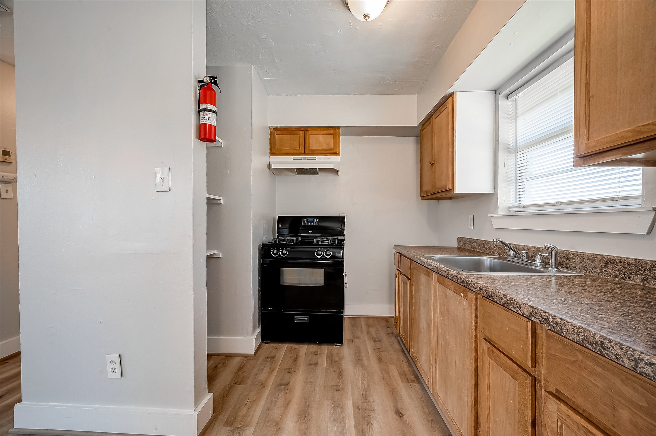 8814 Manus Street, Unit 5 Houston, TX 77093 - Photo 18 of 27 This photo showcases a compact kitchen with a black stove and ample wooden cabinetry. The countertops are a speckled brown, and there's a double sink beneath a window with blinds. The space features light-colored walls and flooring, creating a bright and functional cooking area.