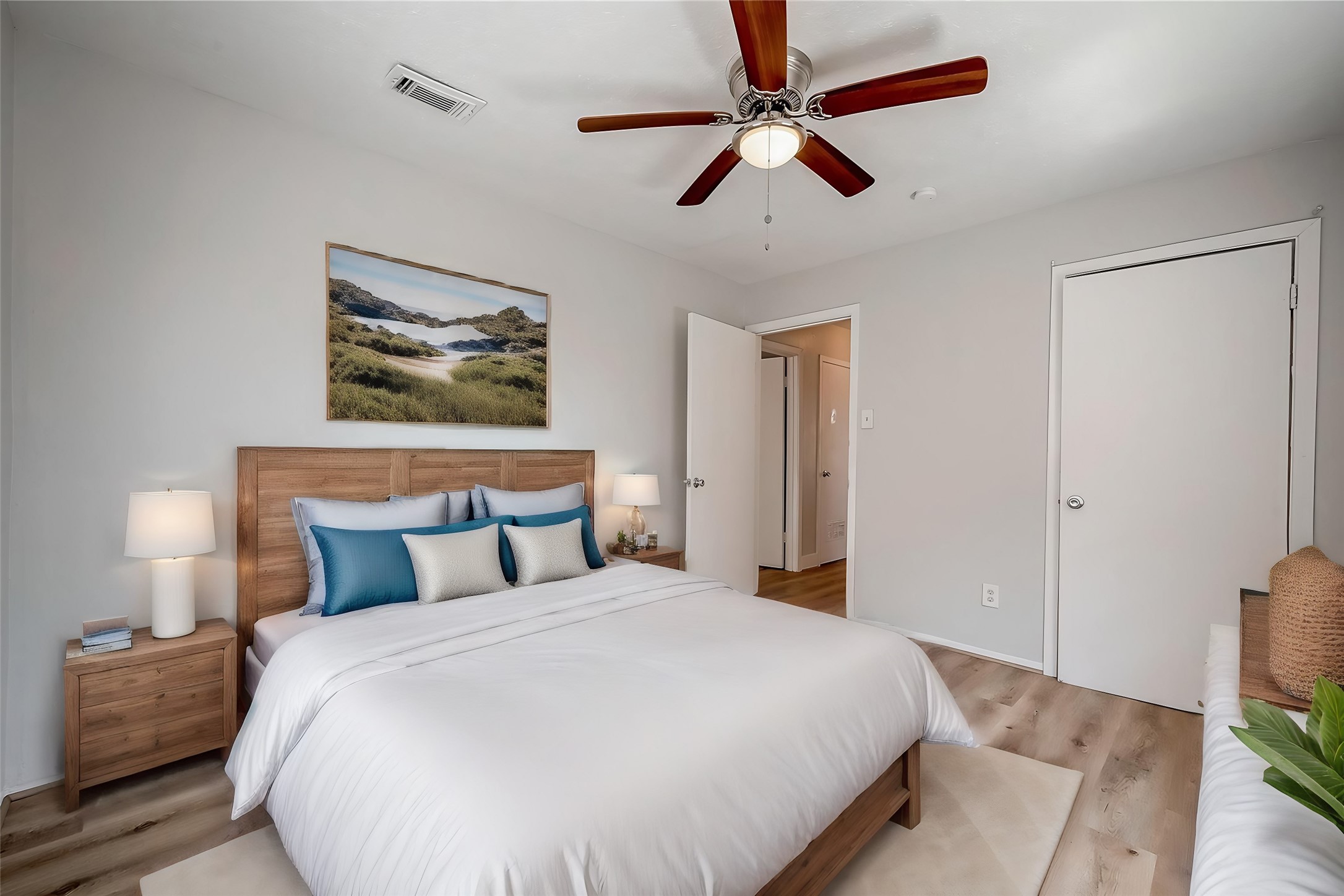 8814 Manus Street, Unit 5 Houston, TX 77093 - Photo 2 of 27 This photo shows a bright, modern bedroom with light-colored walls and wood flooring. It features a double bed with a wooden headboard, matching bedside tables with lamps, and a ceiling fan. The room is decorated with a nature-themed painting, creating a cozy and inviting atmosphere.
