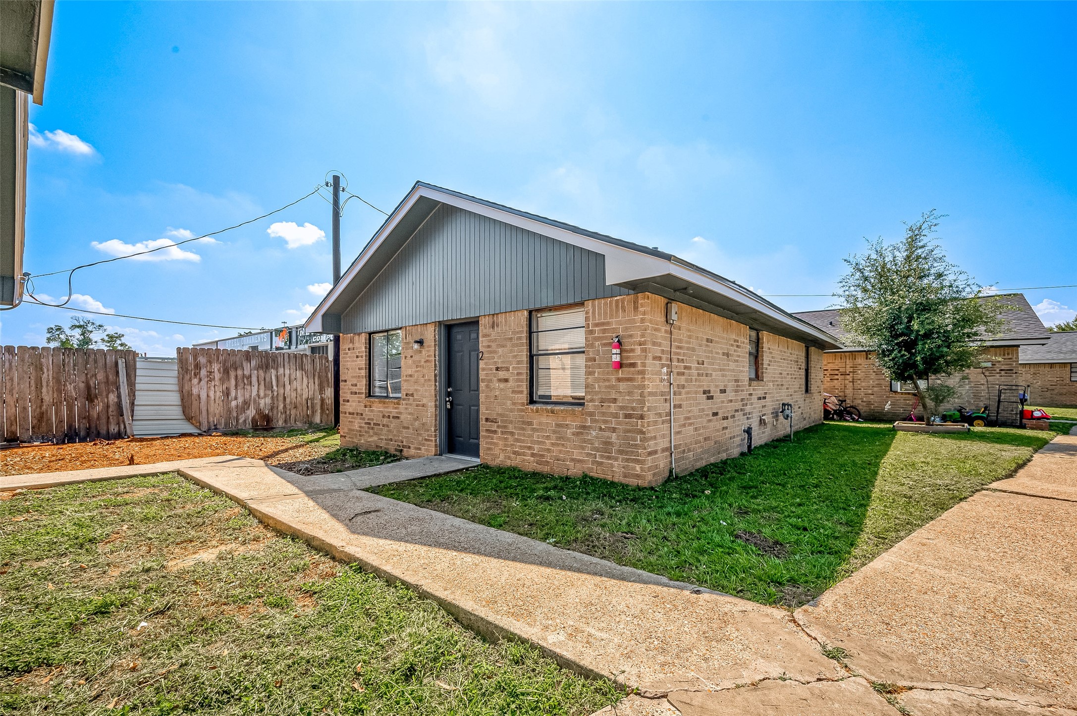8814 Manus Street, Unit 5 Houston, TX 77093 - Photo 4 of 27 This photo shows a single-story, brick home with a sloped roof. The exterior features a small front lawn and a paved walkway leading to the entrance. A wooden fence encloses the side yard, and there's a tree visible in the background. The property appears well-maintained and is situated in a sunny area.