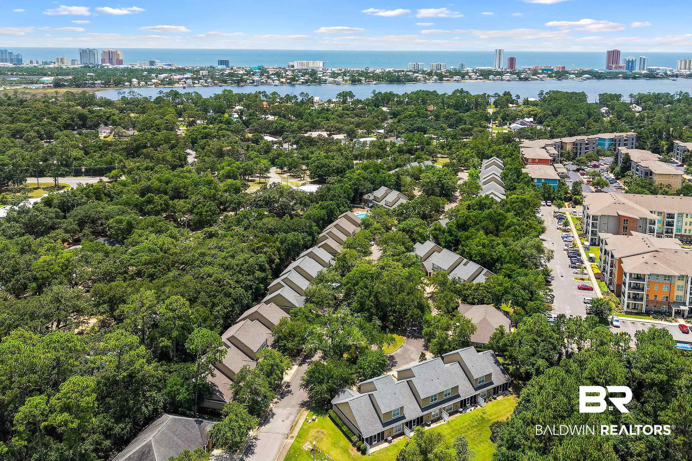 430 West Fort Morgan Road, Unit 704 Gulf Shores, AL 36542 - Photo 33 of 35 Aerial overview revealing an established community with lush tree canopies, residential buildings, and a distant coastline with urban structures
