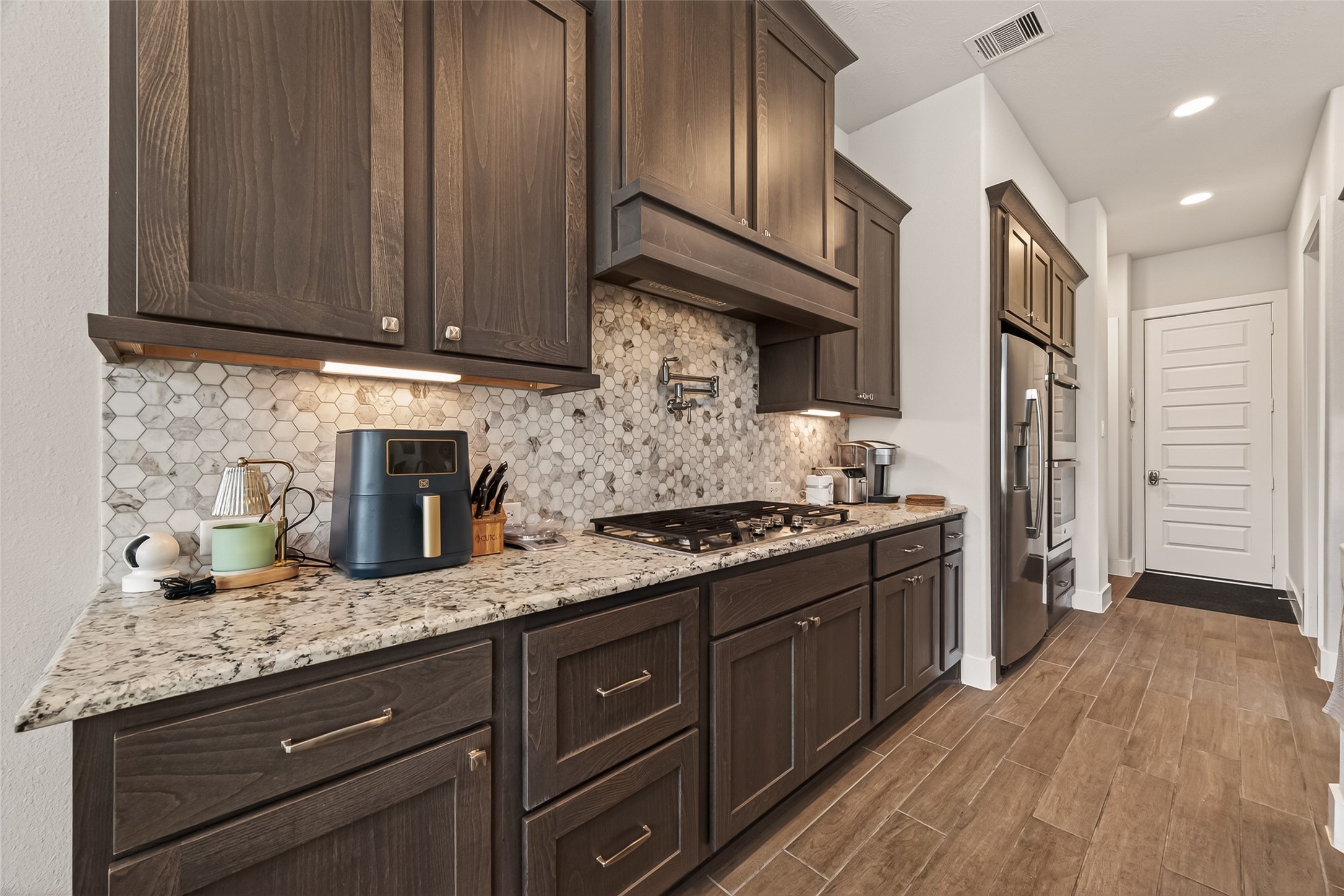 1815 Brea Rdg Trail Katy, TX 77494 - Photo 13 of 43 This kitchen features dark wood cabinetry, granite countertops, and a stylish hexagonal tile backsplash. It includes modern stainless steel appliances, a built-in gas cooktop, and ample storage, creating a sleek and functional cooking space.