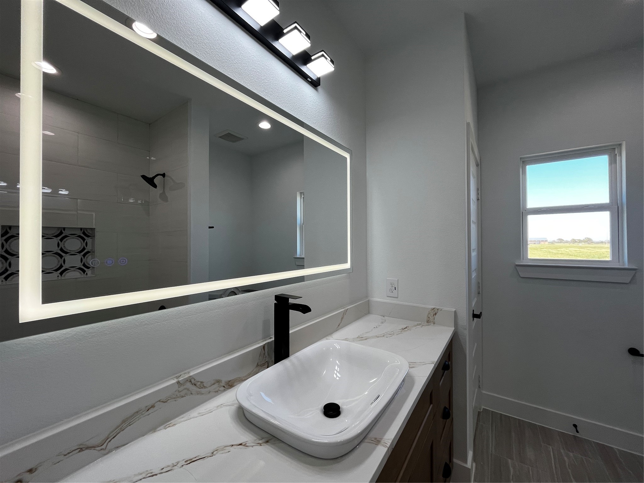 471 Buffalo Trail Angleton, TX 77515 - Photo 21 of 35 Modern bathroom with a sleek design featuring a large illuminated mirror, stylish black fixtures, and a vessel sink on a quartz countertop. The space is brightened by natural light from a window with a scenic view.