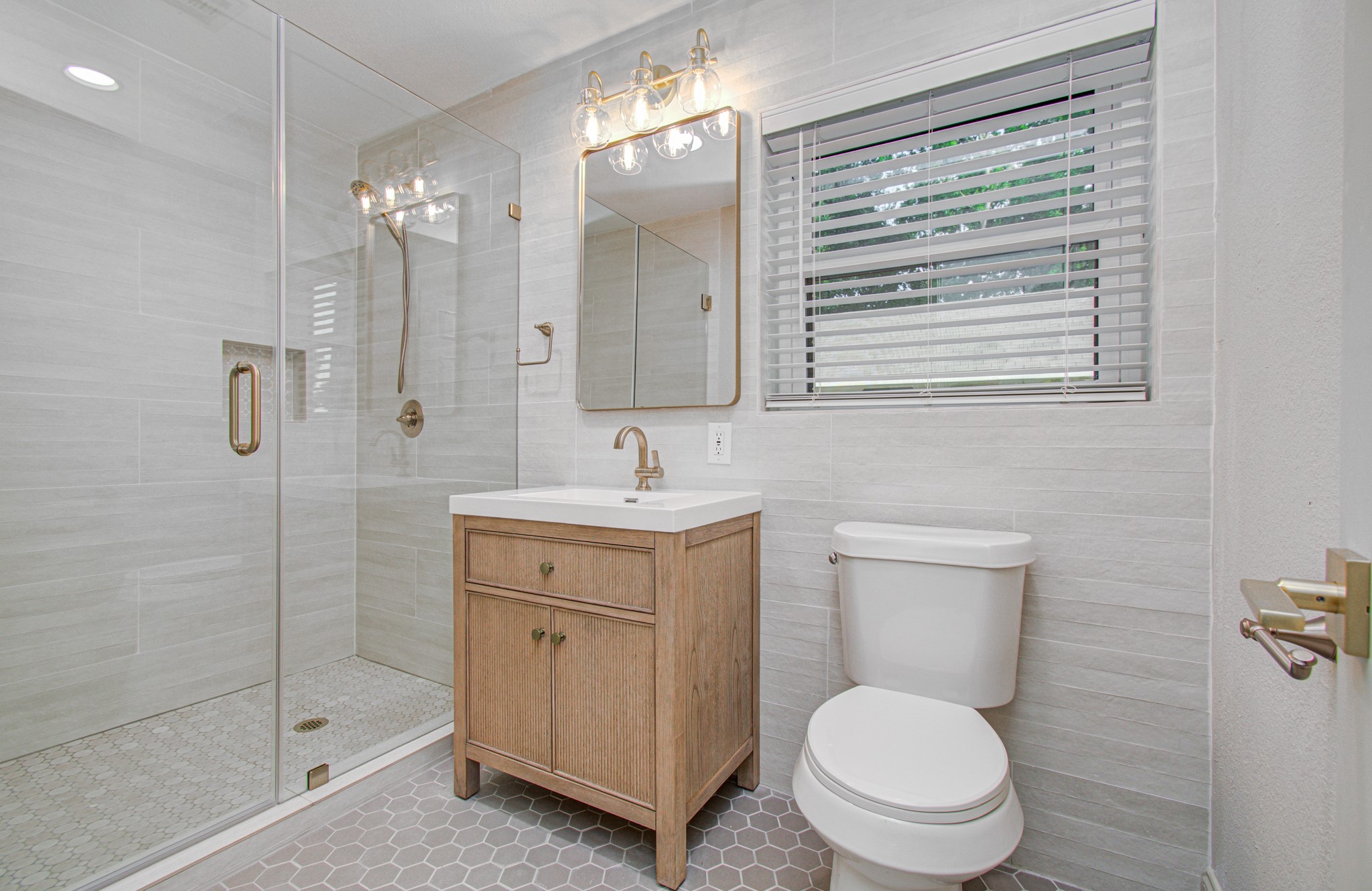 1801 Benbow Way Houston, TX 77080 - Photo 15 of 32 This modern bathroom features a spacious glass-enclosed shower, a sleek vanity with a wood finish, and a contemporary light fixture. Natural light streams through a window with blinds, highlighting the elegant tile work and neutral color palette.