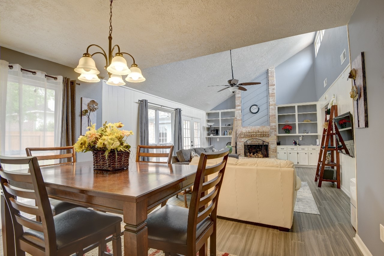 25810 Fairbrook Lane Spring, TX 77373 - Photo 12 of 33 This inviting open-concept living and dining area features a high vaulted ceiling, a cozy brick fireplace, and built-in shelving. Natural light streams through large windows, illuminating the space, while a chandelier adds a touch of elegance over the dining table.