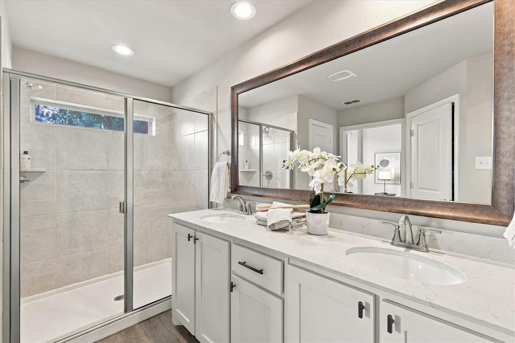 915 Timber Ridge Drive Justin, TX 76247 - Photo 18 of 34 Dual vanity with white cabinetry and integrated sinks, a large framed mirror, and a glass-enclosed shower with tiled walls