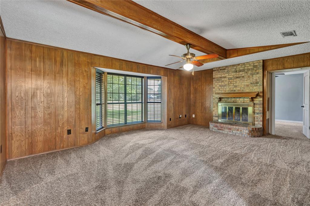 108 Prairie Creek Road Red Oak, TX 75154 - Photo 3 of 19 Living area featuring wood paneling, a brick fireplace with a wood mantle, a bay window with blinds, wood-finish ceiling beams, and a ceiling fan