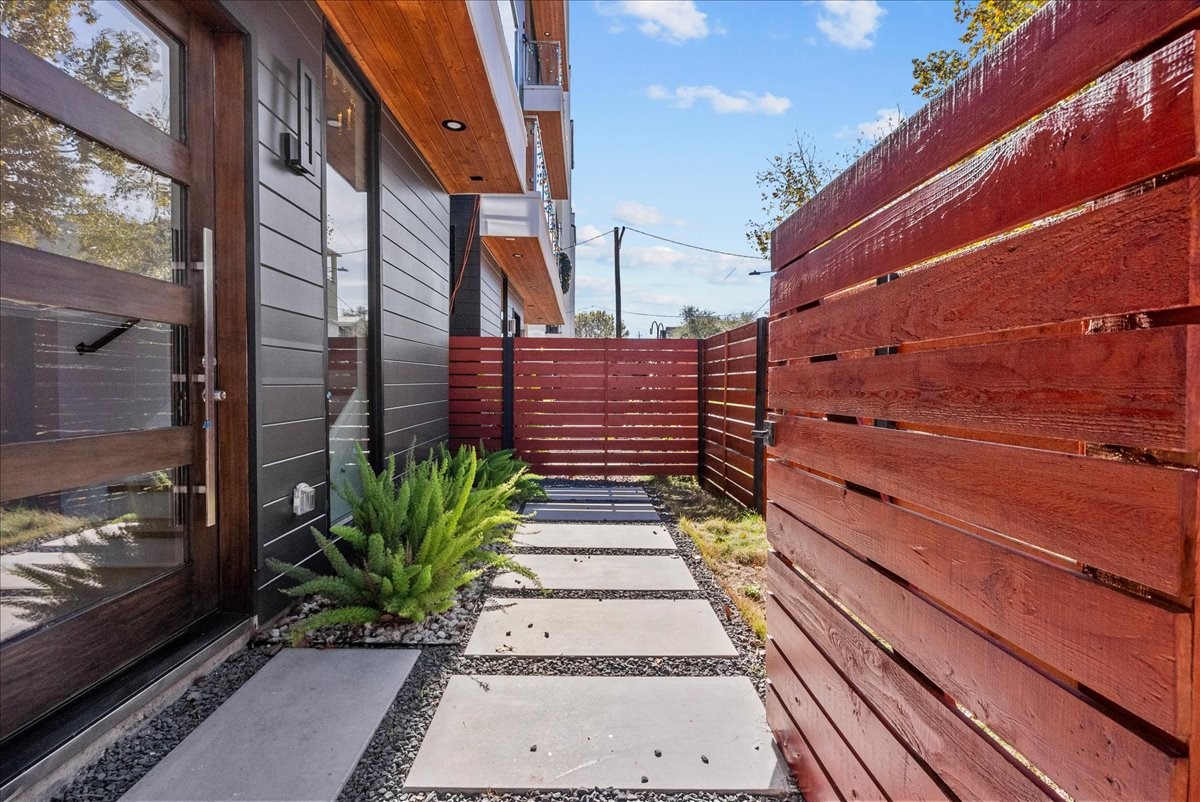 516 Reinerman Street Houston, TX 77007 - Photo 4 of 42 This photo showcases a modern side yard with a sleek wooden fence and a pathway of concrete slabs surrounded by gravel. The exterior features dark siding with a glass door and a small garden area with lush greenery, offering a clean and contemporary outdoor space.