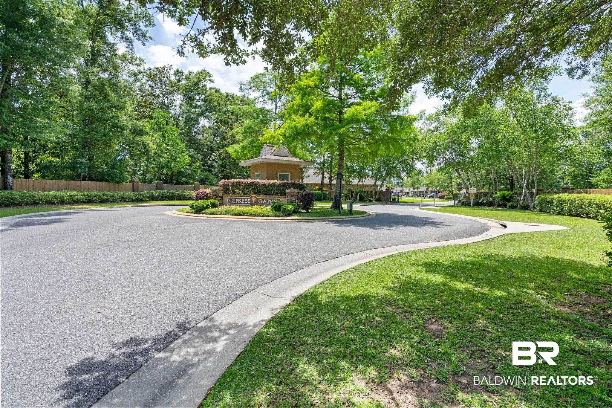 1270 Surrey Loop Foley, AL 36535 - Photo 25 of 27 Paved entry road with a curbed island featuring a stone monument sign, mature trees, manicured green spaces, and a gatehouse structure