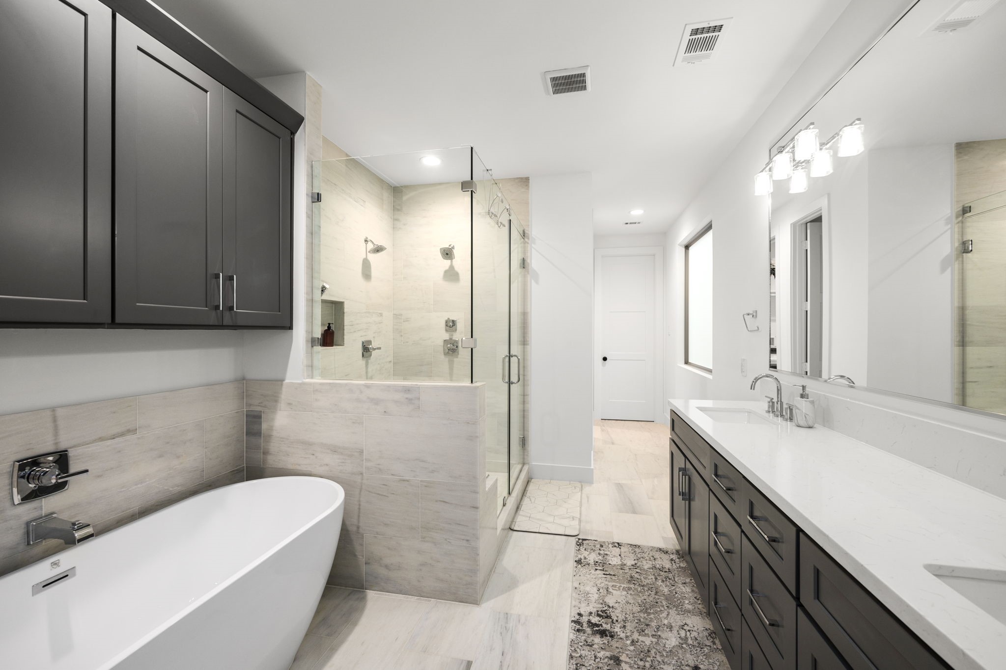 2411 Hazard Street Houston, TX 77019 - Photo 31 of 46 This additional view of the primary bathroom highlights the sleek freestanding tub and a spacious glass-enclosed shower with dual shower heads and built-in shelving. The large vanity offers ample storage space with modern dark cabinetry and polished finishes. The combination of clean lines, neutral tones, and luxurious fixtures adds a contemporary feel to the space.