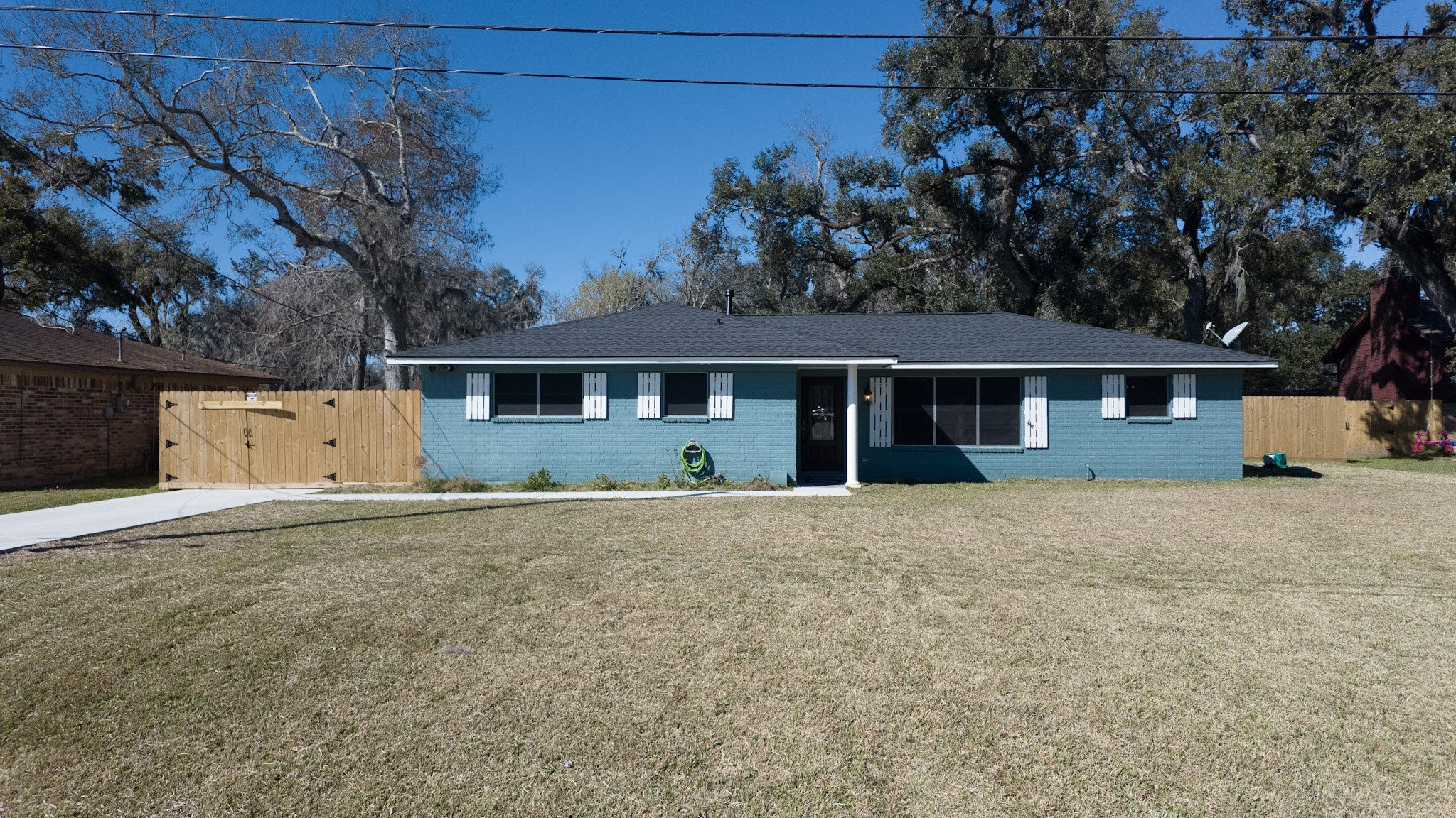 This photo shows a charming single-story home with a blue exterior and white trim. It features a spacious front yard and a neat driveway, with a wooden fence enclosing the property. Mature trees provide a scenic backdrop.