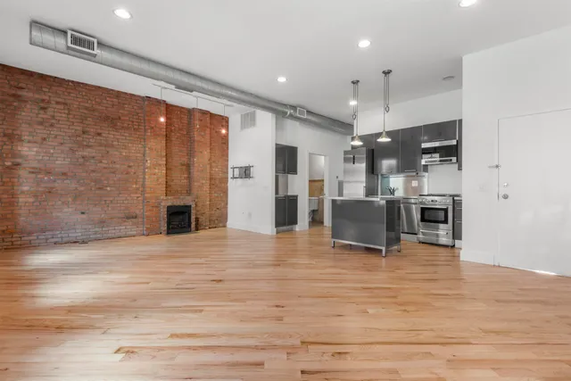 a view of an empty room and kitchen with stainless steel appliances wooden floor