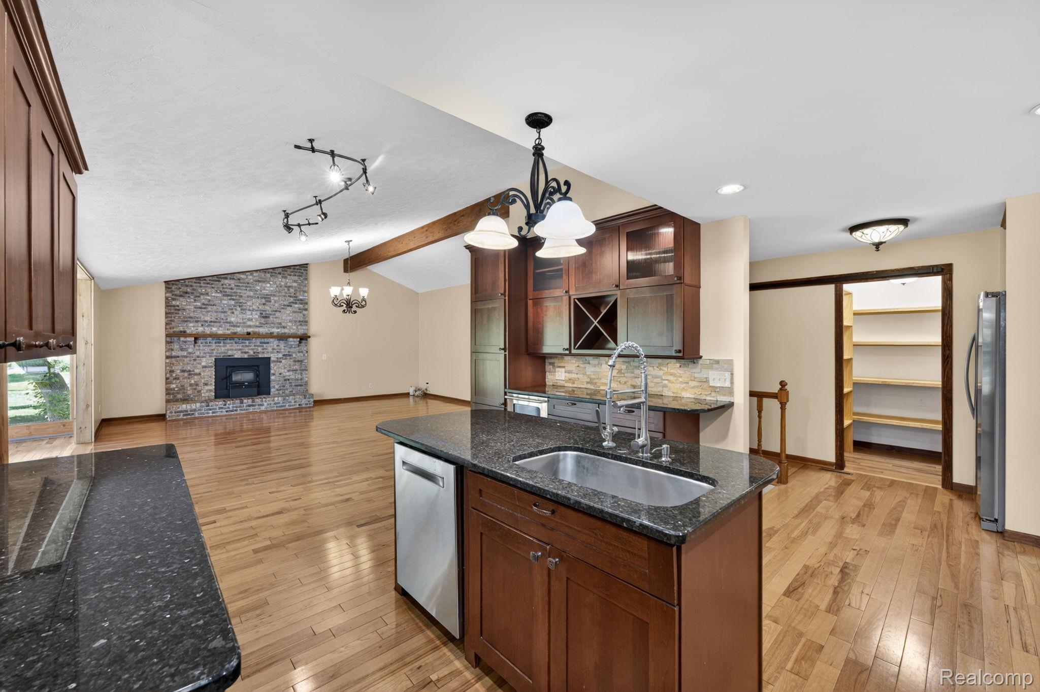 9353 Hill Road Swartz Creek, MI 48473 - Photo 23 of 98 Open-concept living space featuring a granite-topped kitchen island with an undermount sink, hardwood-finish flooring, a stone-faced fireplace, and vaulted ceilings with exposed wood beams