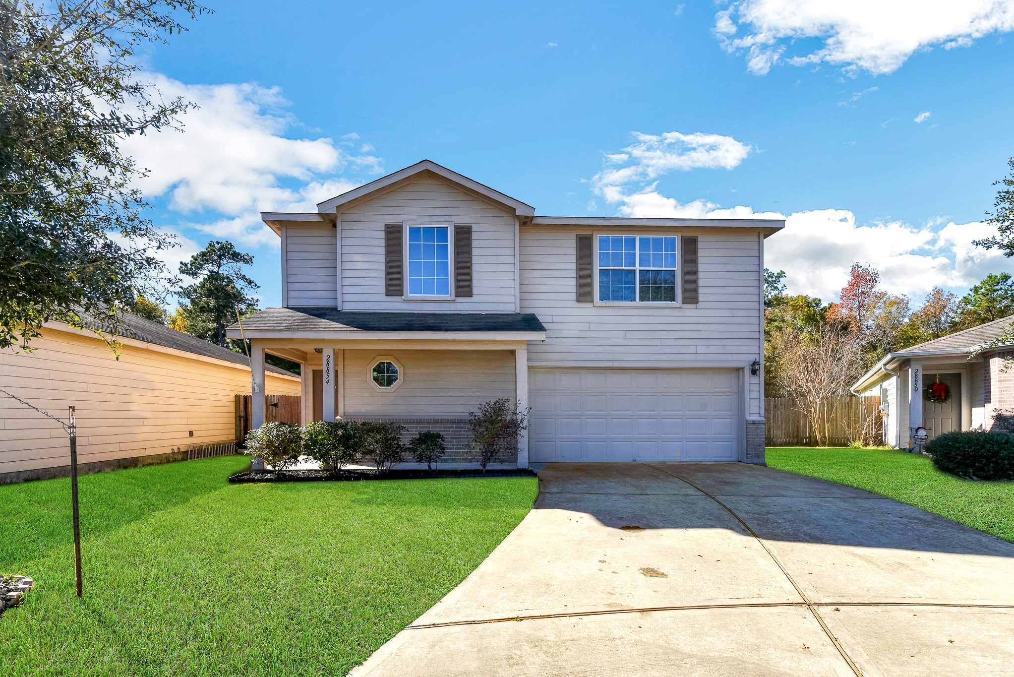 This photo showcases a two-story suburban home with a neatly manicured lawn. The house features a double garage, light siding, and large front windows. It's situated in a quiet neighborhood with mature trees in the background.