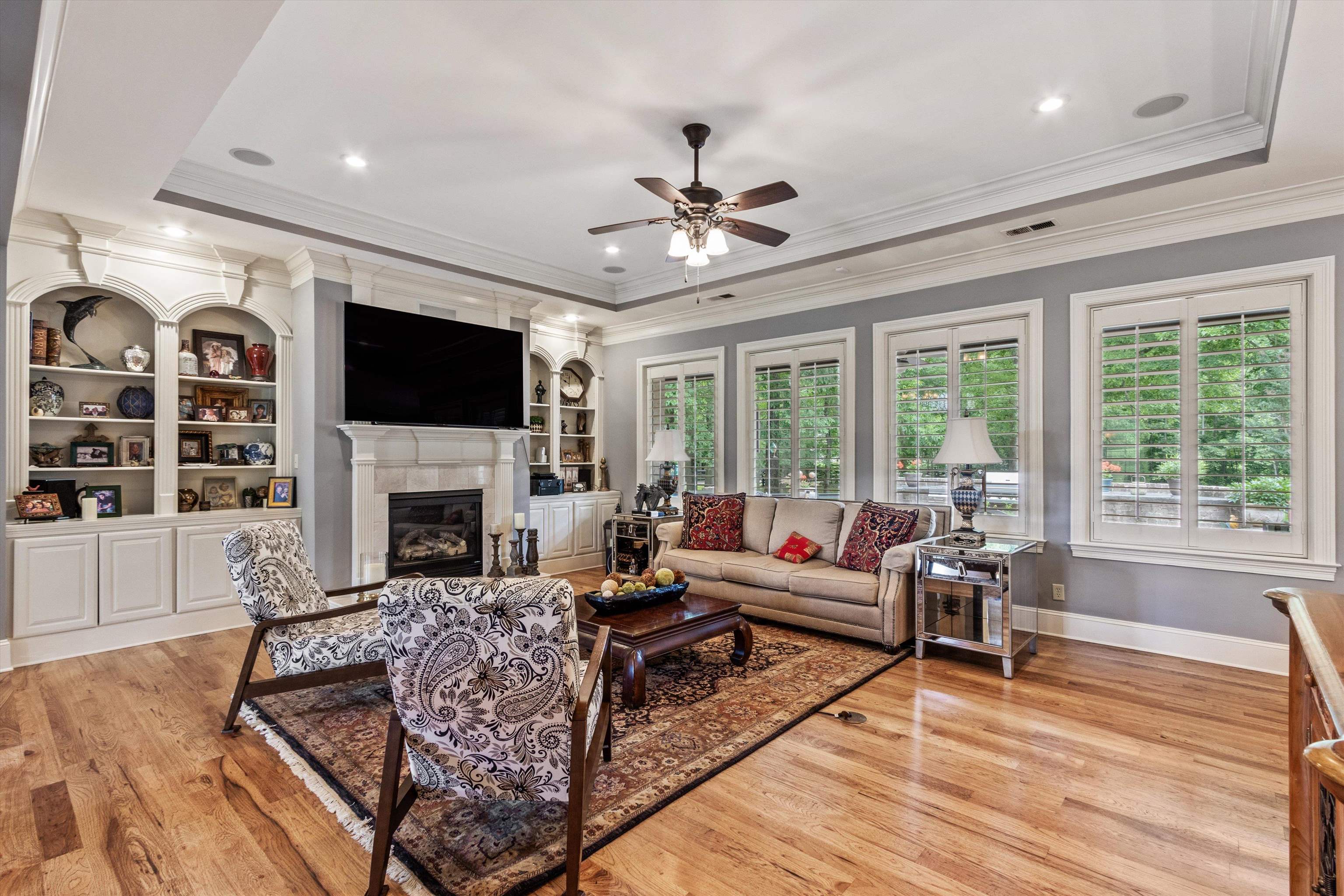 150 Ridgecrest Drive Arlington, TN 38002 - Photo 12 of 40 Living area featuring hardwood flooring, a gas fireplace with a stone surround, built-in shelving, a tray ceiling with recessed lighting, and multiple windows with plantation shutters