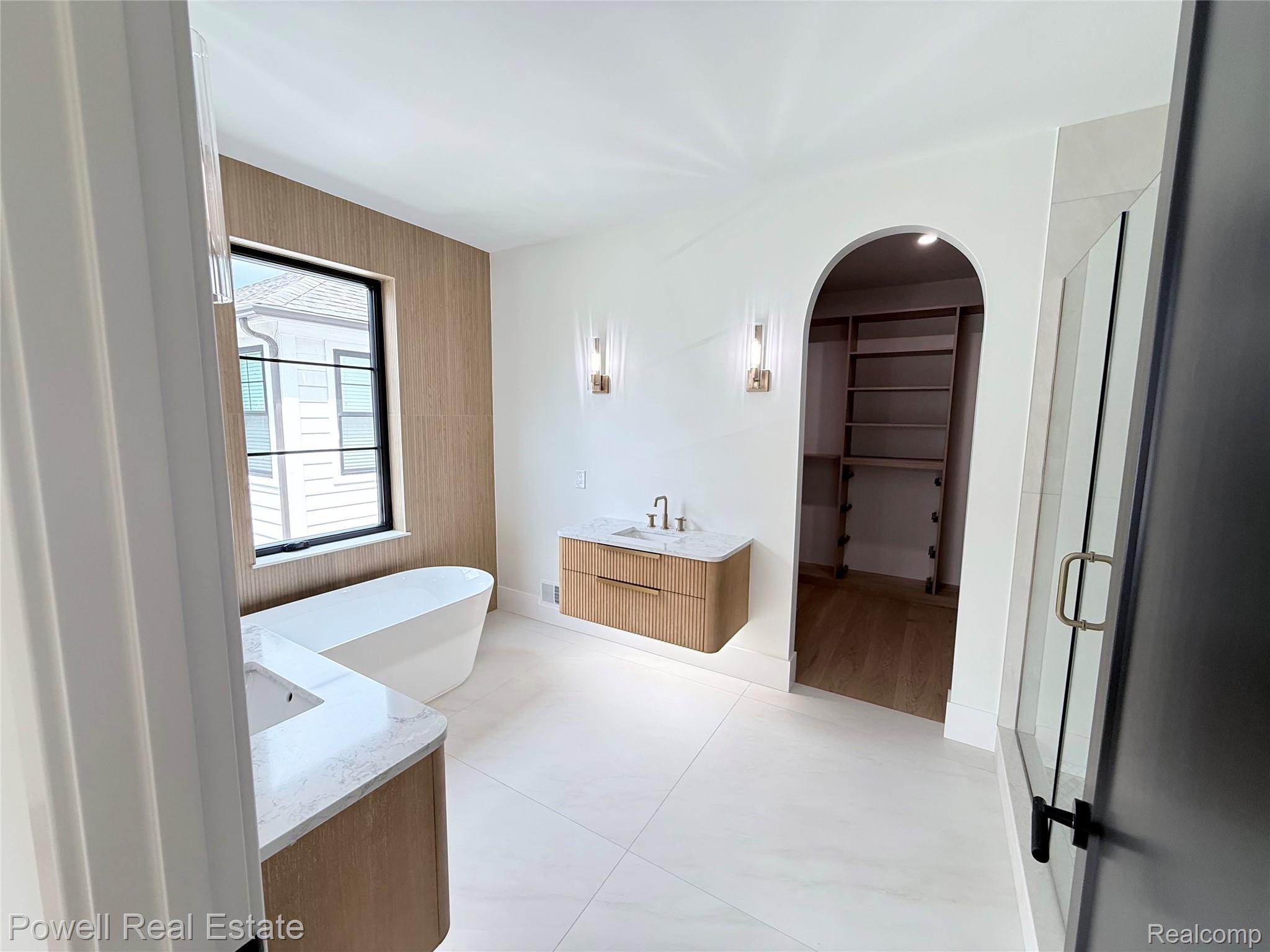 1998 Hazel Street Birmingham, MI 48009 - Photo 14 of 22 Spacious bathroom featuring a freestanding soaking tub, wood-finish wall paneling, dual floating vanities with stone countertops, and an arched entry to a custom closet with built-in shelving