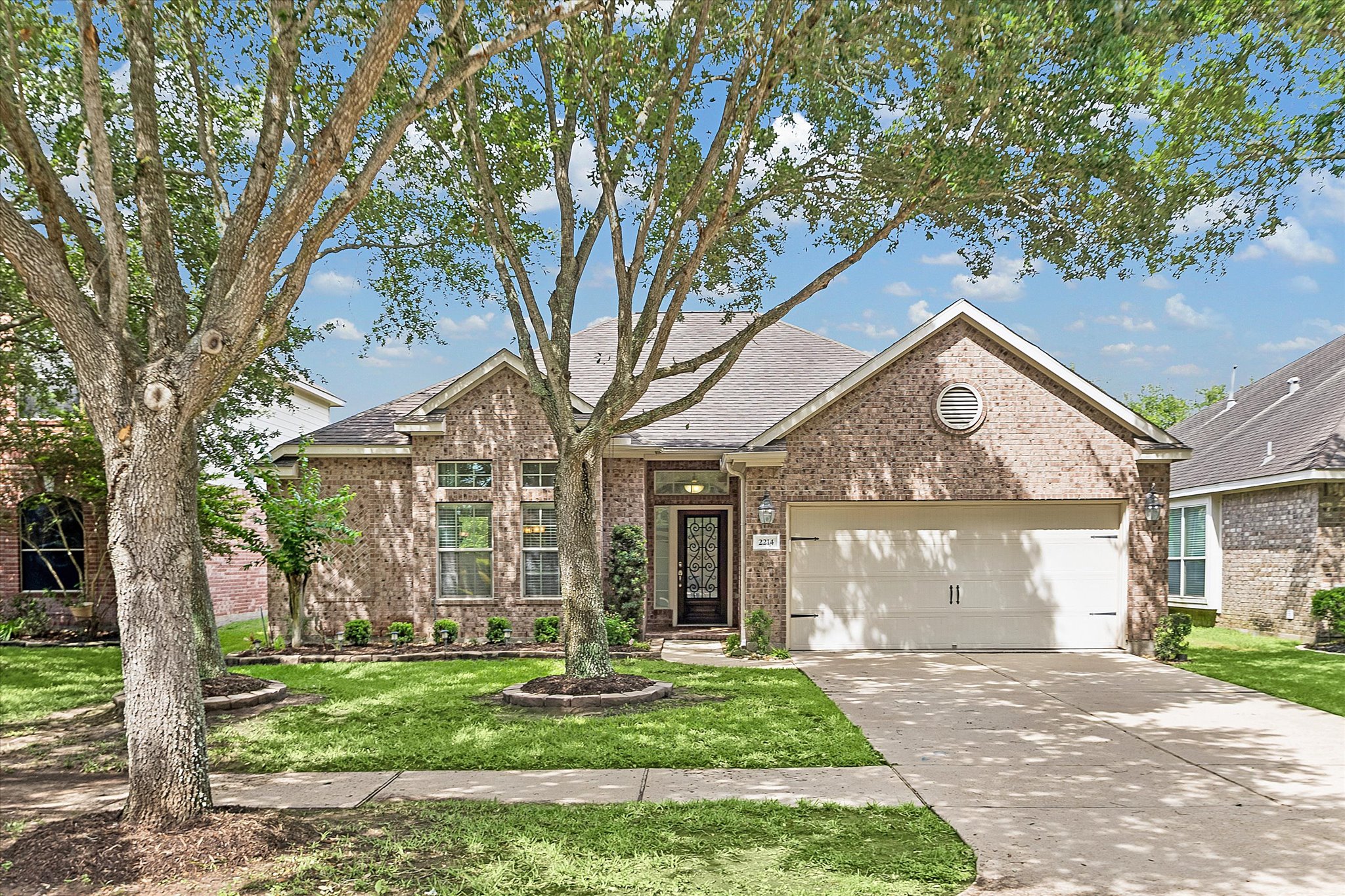 Brick-veneer exterior featuring an attached two-car garage, mature trees, and a concrete driveway