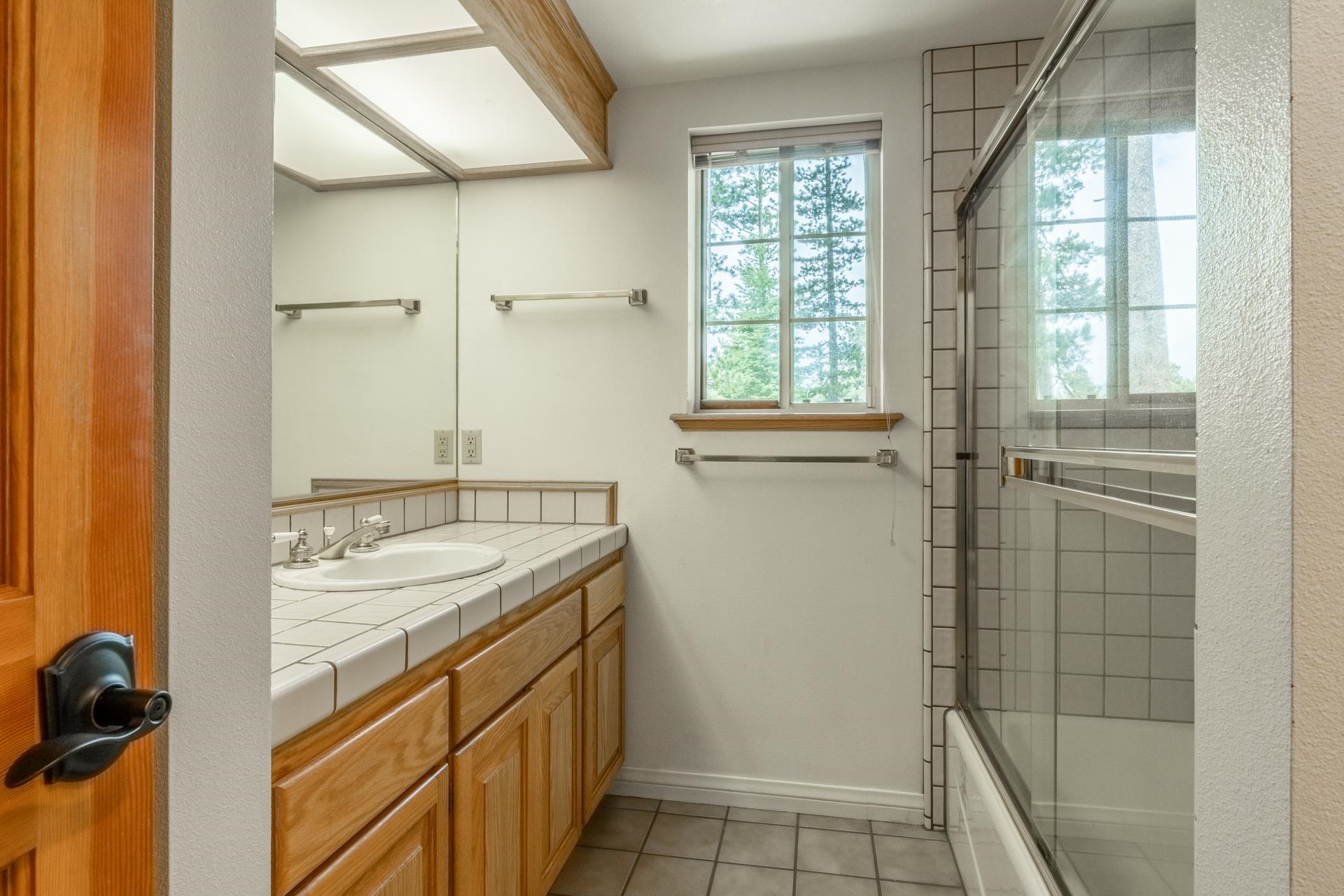 272 Ridge Way Mammoth Lakes, CA 93546 - Photo 40 of 55 Bathroom featuring a wood-finish vanity with a tile countertop, a full-width wall mirror, and a shower-tub combination with a glass enclosure