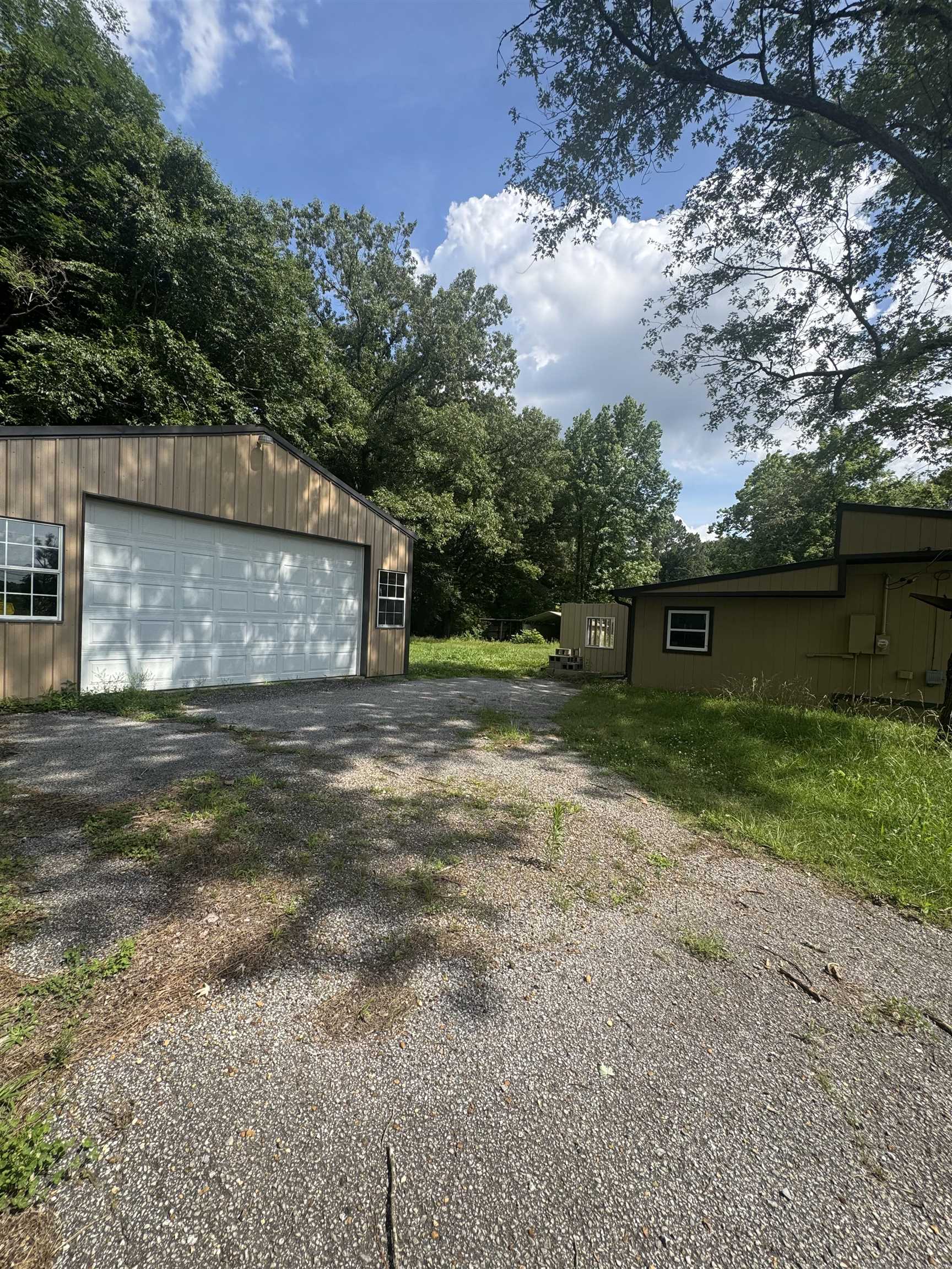 106 Cole Road Drummonds, TN 38023 - Photo 9 of 21 Gravel parking area fronting a garage with light-toned siding and multiple windows, surrounded by mature trees and natural landscaping