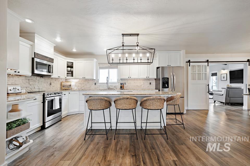 23800 Breyer Road Caldwell, ID 83607 - Photo 11 of 50 Modern kitchen featuring white cabinetry, stainless steel appliances, an oversized island with seating, a brick-patterned backsplash, and wood-finish flooring