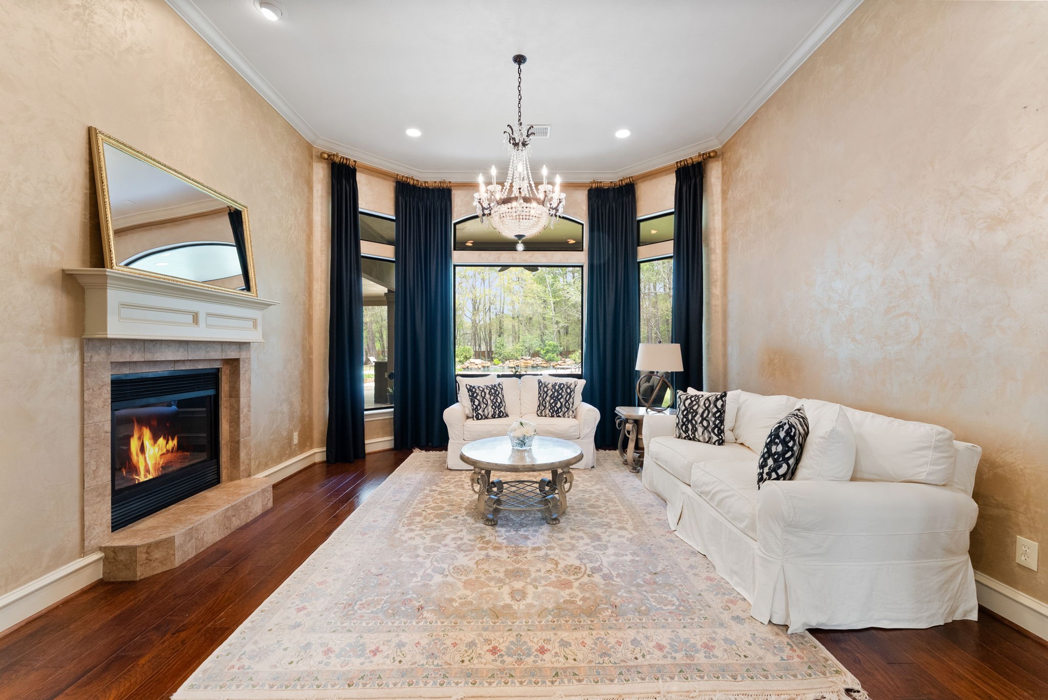 3907 Rayford Road Spring, TX 77386 - Photo 11 of 38 The Formal Living room is filled with natural light from oversized bay windows with transoms and offers beautiful views of the backyard and pool, with wood floors, crown molding, a statement chandelier, and a double-sided gas fireplace with mantle.