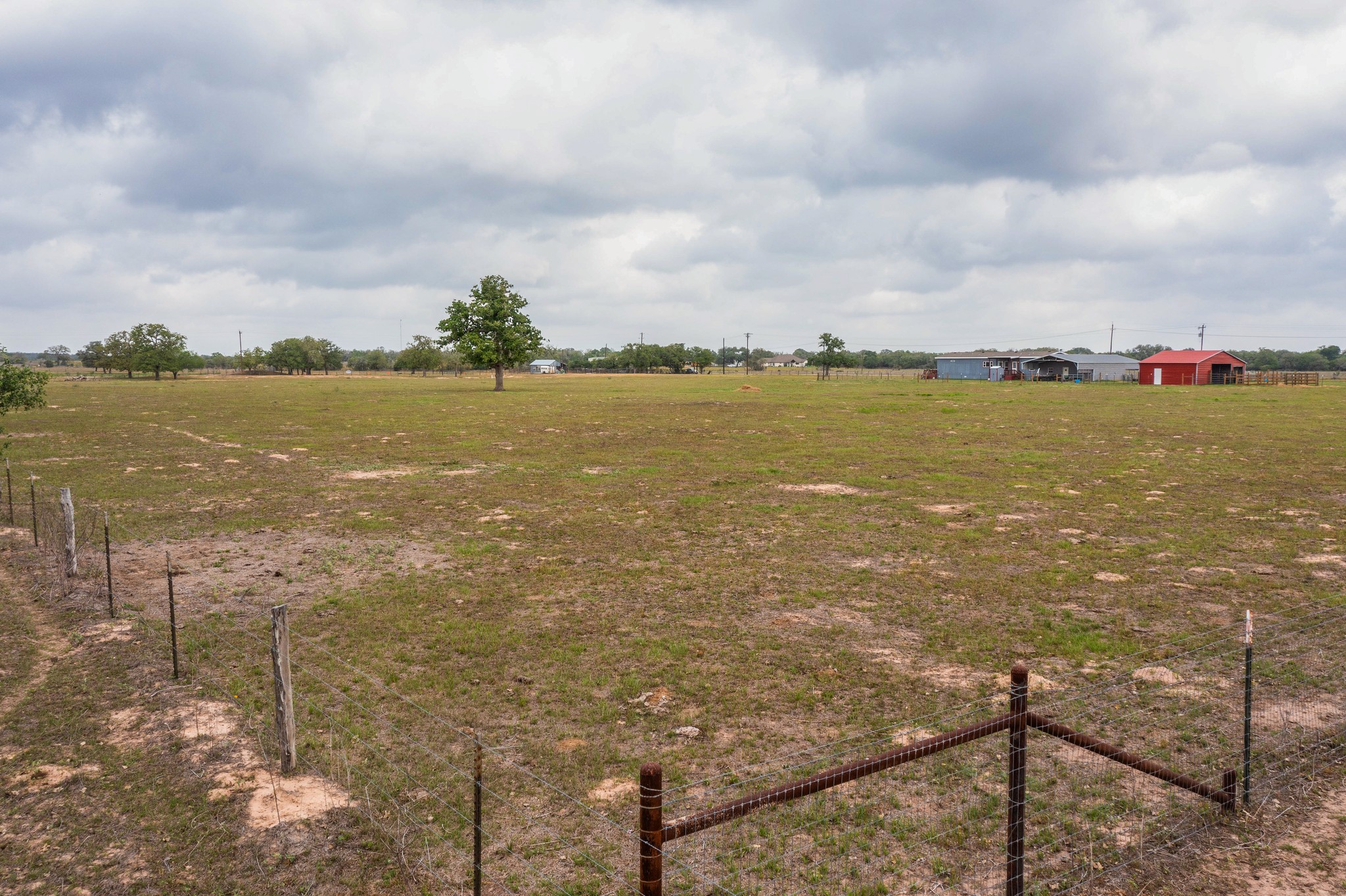 664 Garrett Road Yoakum, TX 77995 - Photo 38 of 39 Expansive open land featuring a wire fence line, scattered mature trees, and distant outbuildings with red and grey roofs