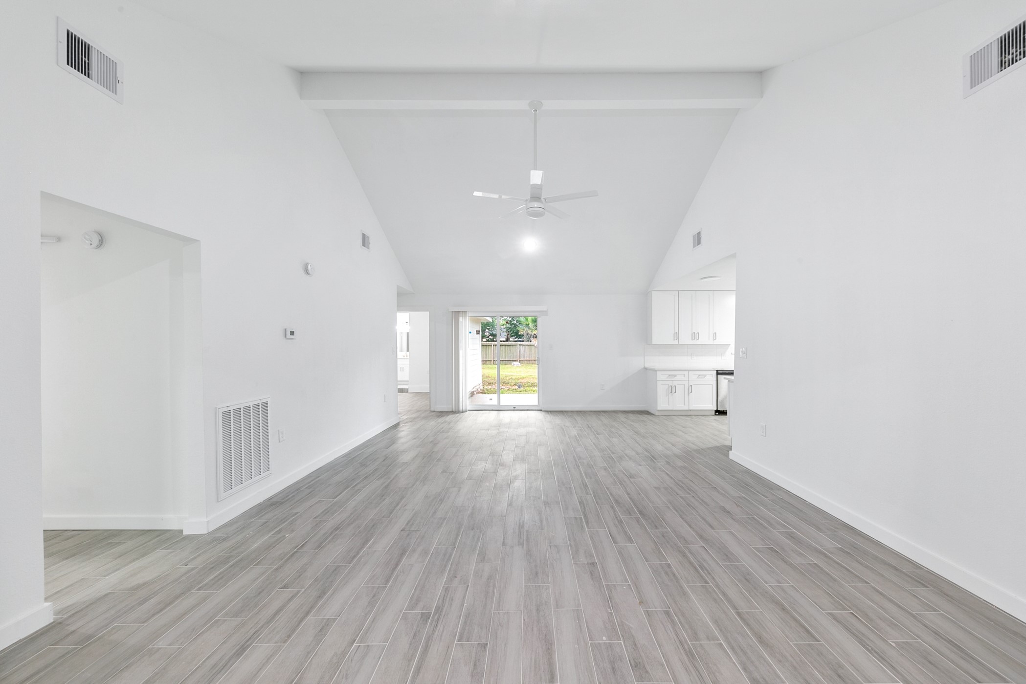 6811 McCullum Road Houston, TX 77489 - Photo 2 of 22 This photo showcases a spacious, open-concept living area with high ceilings and light wood-look flooring. The white walls and ceiling fan contribute to a bright, airy feel, while large sliding doors lead to an outdoor area. The kitchen in the background features white cabinetry, adding to the modern, clean aesthetic.