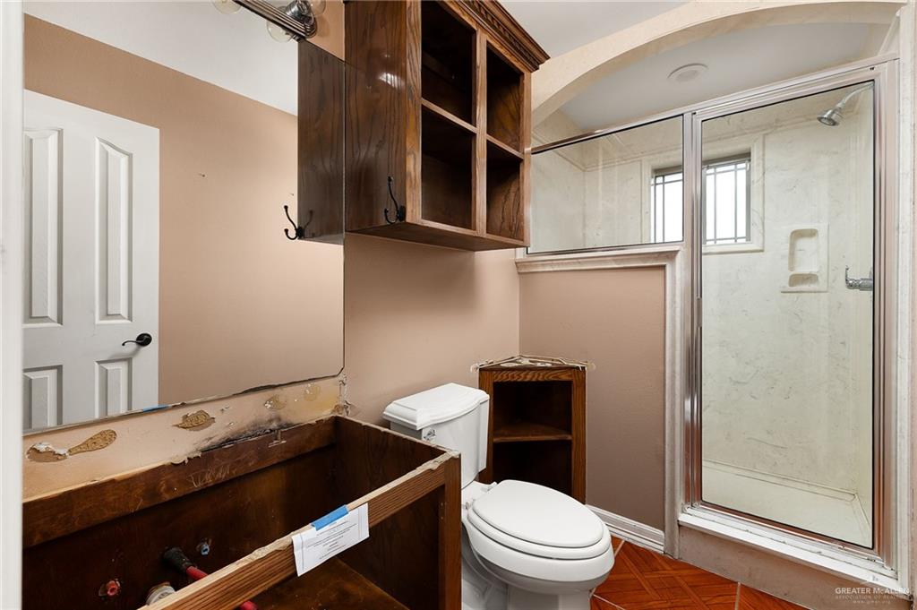 1315 Ruger Street Donna, TX 78537 - Photo 11 of 17 The bathroom features a glass-enclosed shower, a toilet, a wall-mounted wooden cabinet with open shelving, and a mirror