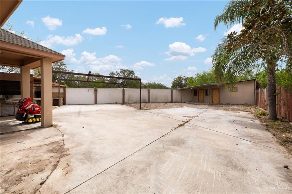 1315 Ruger Street Donna, TX 78537 - Photo 13 of 17 The property features an expansive concrete paved area, a covered patio, and a structure with multiple doors, bordered by a fence and a palm tree