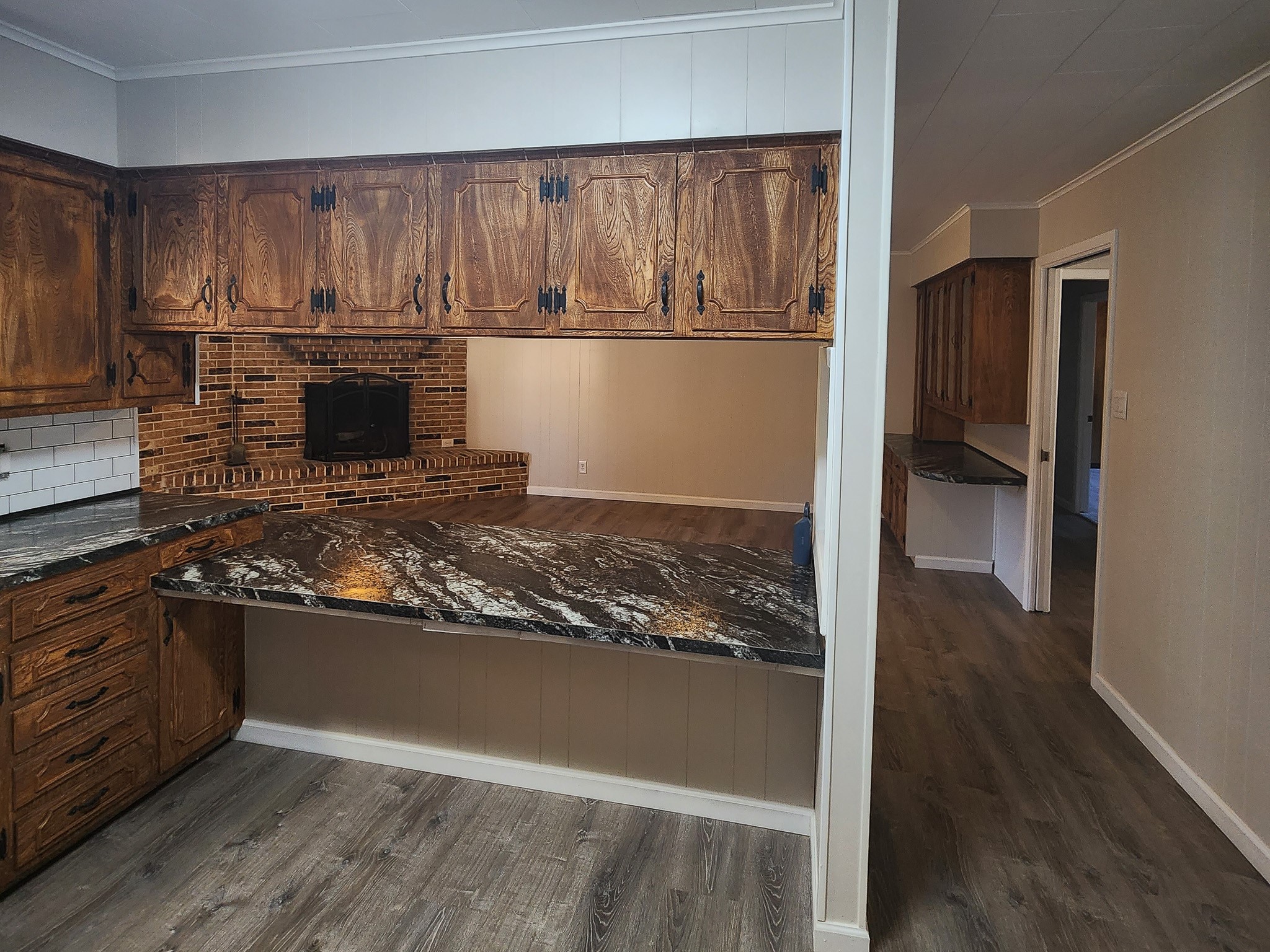 201 Verna Street Jasper, TX 75951 - Photo 11 of 27 This photo showcases a cozy kitchen with rustic wooden cabinets and a dark marble countertop. It features a brick backsplash with a built-in stove, and a spacious layout with an adjacent hallway leading to other rooms. The flooring is a warm, wood-like laminate.