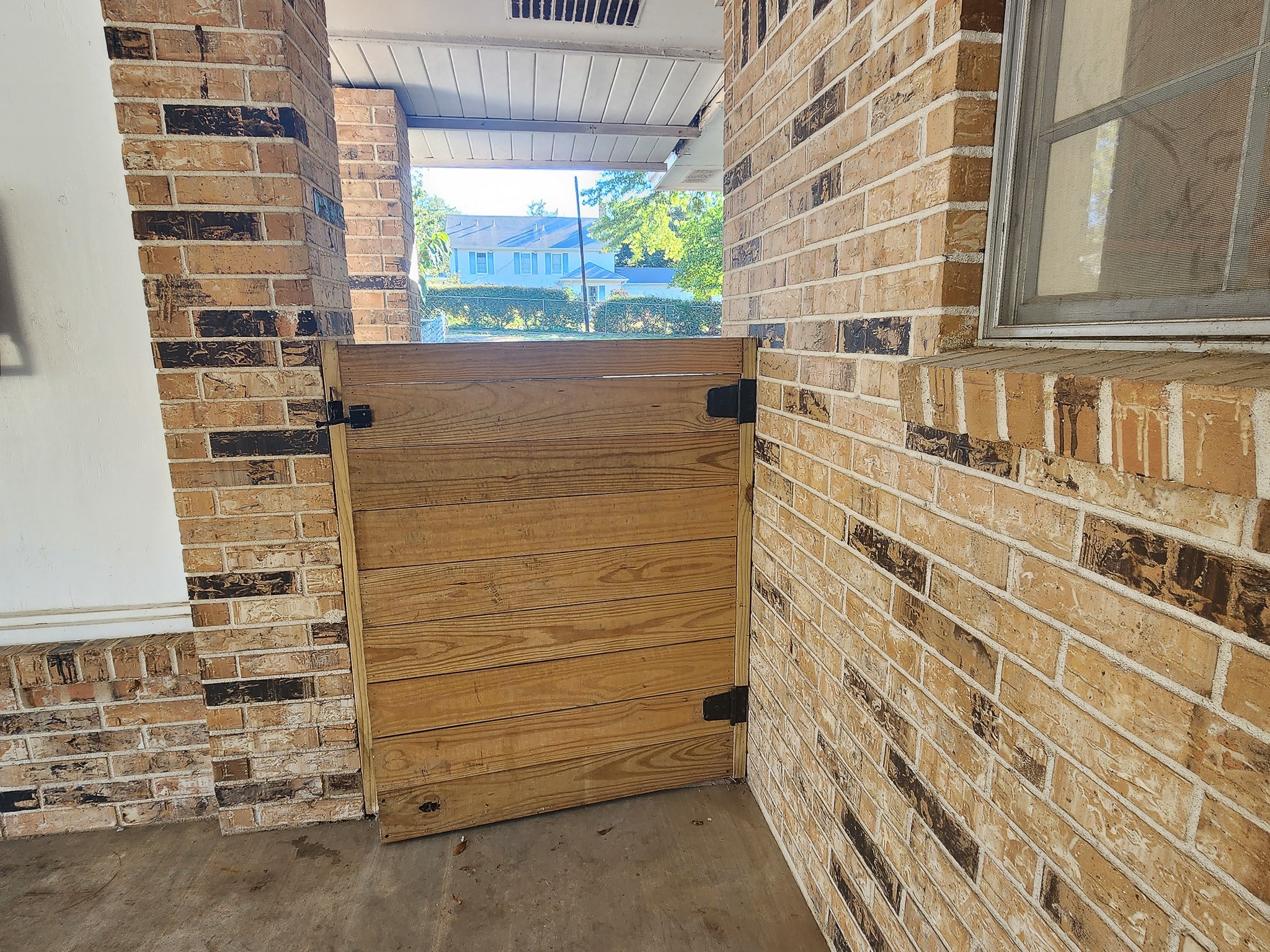 201 Verna Street Jasper, TX 75951 - Photo 20 of 27 This photo shows a brick porch area with a wooden gate. The space has a rustic feel with exposed brick walls and a window on the right, offering a glimpse of greenery outside.