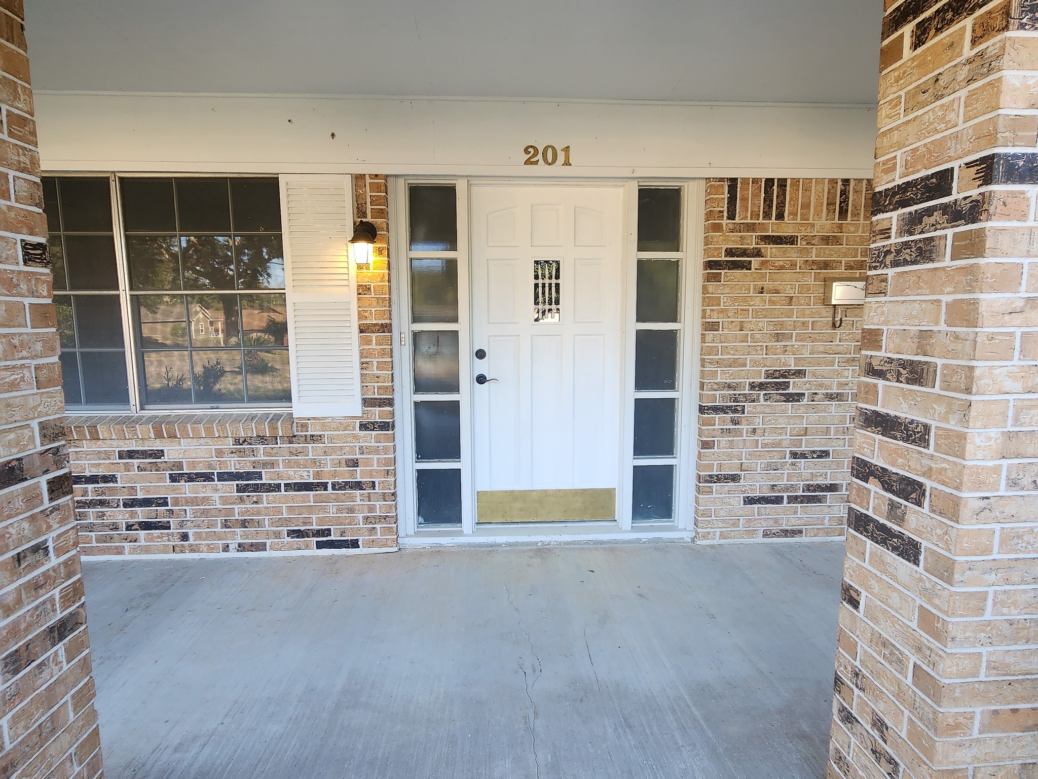 201 Verna Street Jasper, TX 75951 - Photo 21 of 27 This photo shows a welcoming front porch with a white door framed by brick walls and a sheltered entryway. The house number "201" is displayed above the door, which is flanked by glass panels and lit by a wall-mounted light.