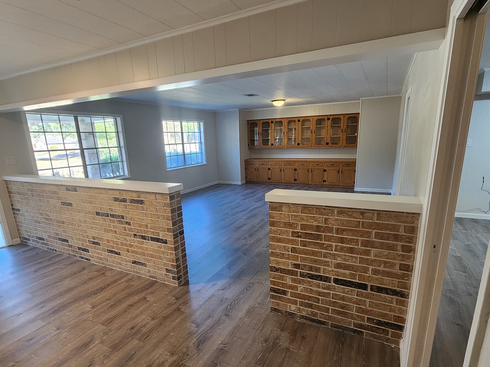 201 Verna Street Jasper, TX 75951 - Photo 3 of 27 This spacious living area with large windows offering ample natural light. It features a partial brick partition and vinyl flooring, leading to a built-in wooden cabinet unit, perfect for storage or display.