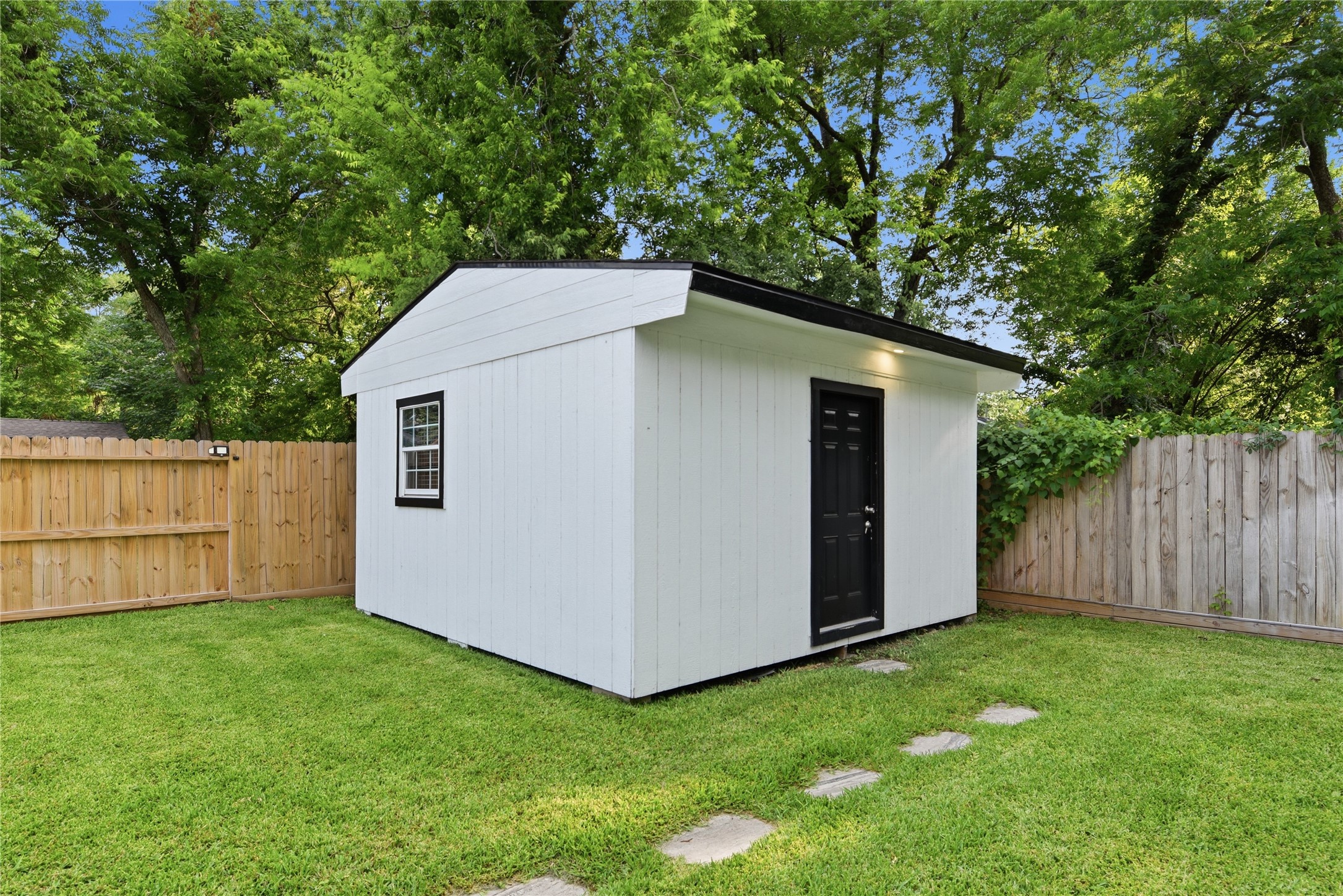 9403 Spaulding Street Houston, TX 77016 - Photo 36 of 50 This photo features a charming, small white shed with a black door and window, set in a well-maintained, fenced backyard with lush green grass and mature trees, offering a serene outdoor space.